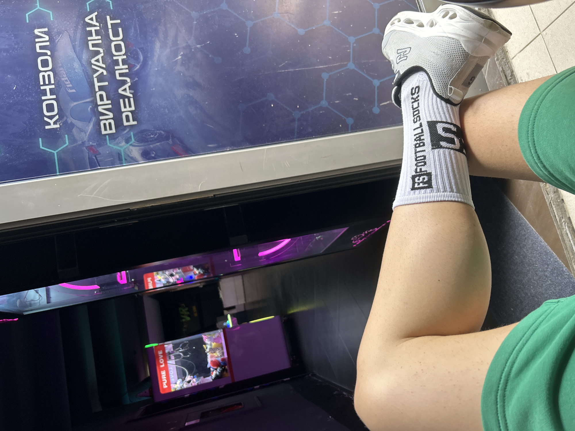 The image shows a person's legs, wearing a green shorts and white socks with black text that reads "FS FOOTBALLSOCKS". The person is wearing a grey sneaker. The background appears to be an arcade or entertainment center. There are arcade machines with neon lights visible, including a "PURE LOVE" claw machine and a VR-themed area. A large digital display with text in Cyrillic script, possibly advertising "КОНЗОЛИ" (consoles) and "ВИРТУАЛНА РЕАЛНОСТ" (virtual reality), is also visible. The floor is tiled and dark. The scene suggests an indoor entertainment venue, possibly in the city of Burgas, Bulgaria, based on the Cyrillic text.