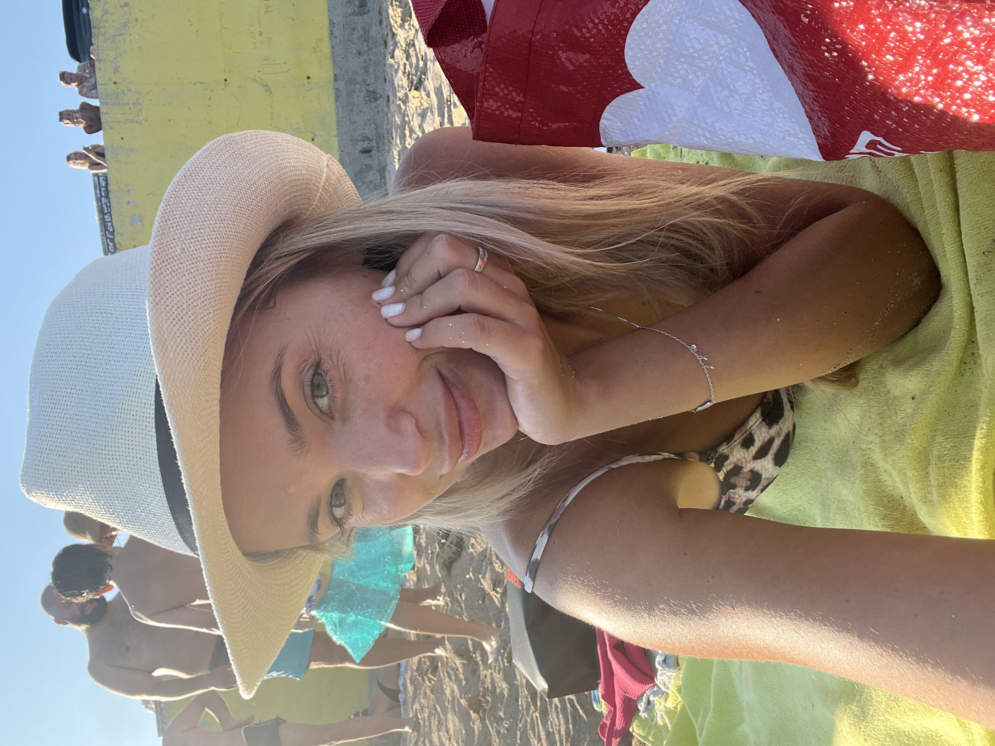 A blonde woman wearing a white straw hat and a leopard-print bikini lies on a yellow towel on a sandy beach, her hand resting on her cheek. In the background, other beachgoers are visible, including a man with a beard and a person in a blue tutu. A large red and white bag is to the right of the woman. The scene suggests a sunny day at the beach in Fontanka, Ukraine.