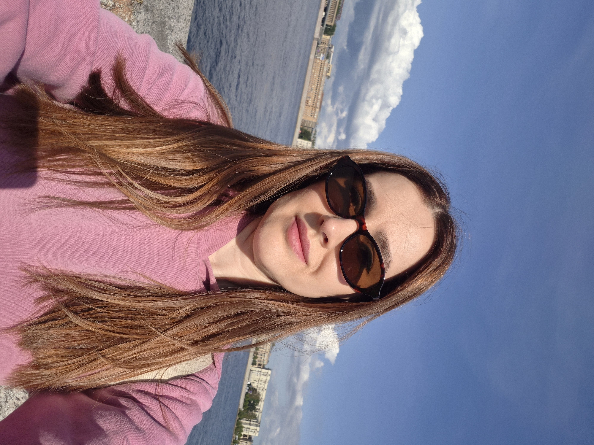 A woman with long, reddish-brown hair and sunglasses is taking a selfie by the Neva River in Saint Petersburg, Russia. She is wearing a pink hoodie. In the background, there are buildings along the river and white clouds in a blue sky.