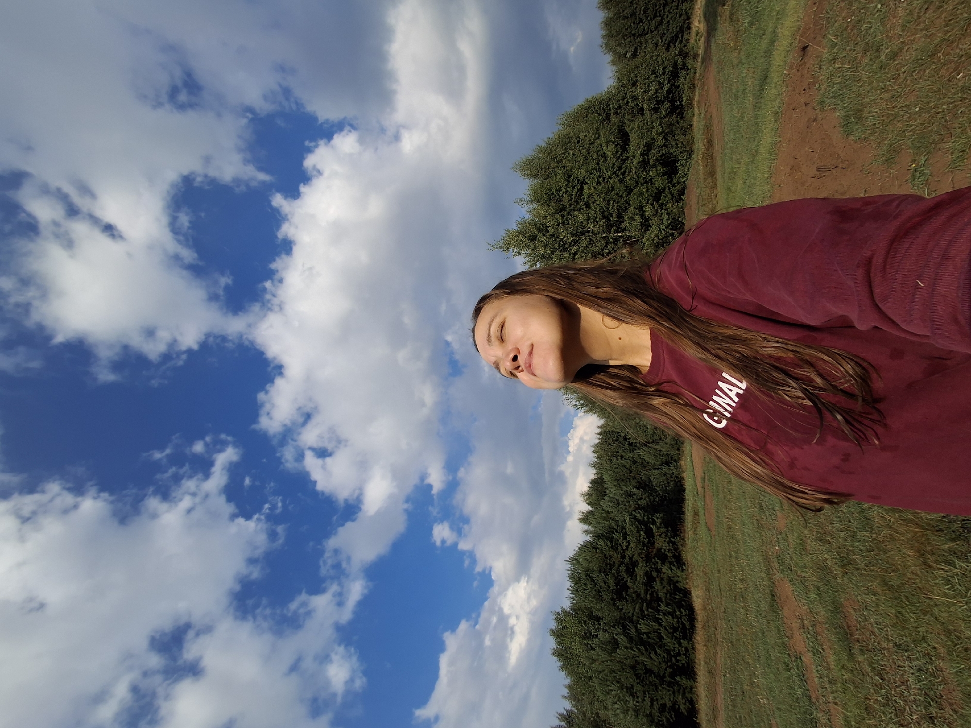 In Plishkino, Russia, a young woman stands in a grassy field, looking up at a bright blue sky filled with white, puffy clouds. She has long, brown hair and is wearing a maroon sweatshirt with white lettering that reads "GINAL." The field is bordered by a dense line of green trees. The sun is casting a warm glow on her face, and she appears to be enjoying the moment. The overall scene suggests a peaceful and natural setting.