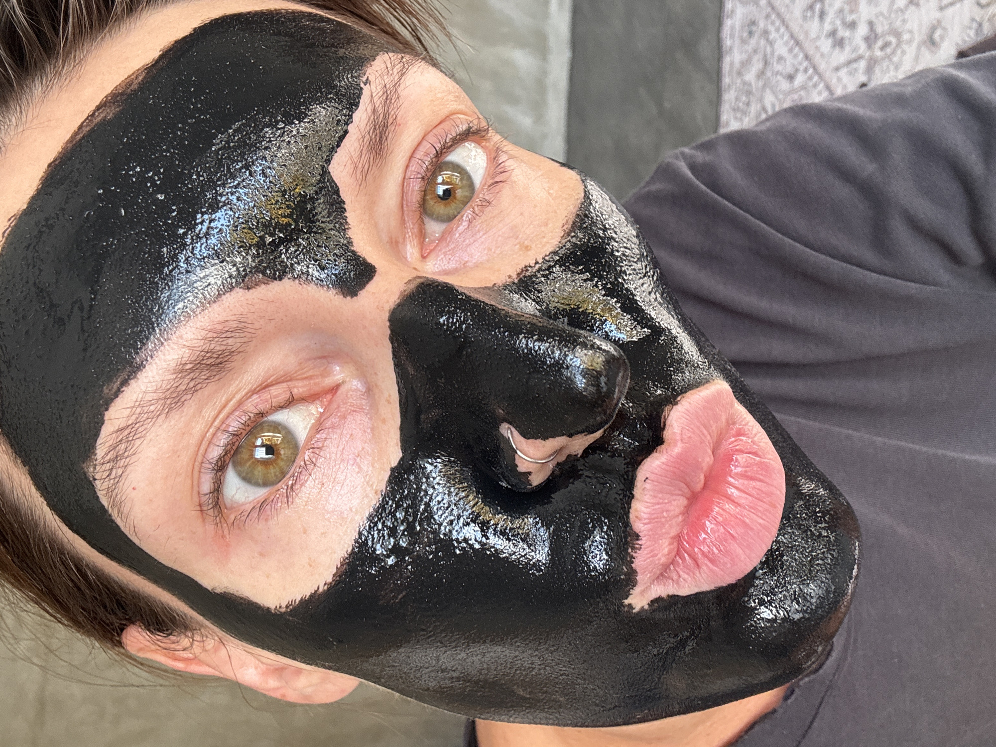 A close-up image shows a person's face covered in a black charcoal mask, with only their eyes, nose, and lips visible. The person has light skin and brown eyes, with a silver hoop piercing on their left nostril. The mask is thick and glossy, reflecting light. The person is wearing a dark grey t-shirt. The background is out of focus and appears to be indoors, with a textured wall and a patterned rug visible. The image does not contain information about the city of North Kuta, Indonesia.