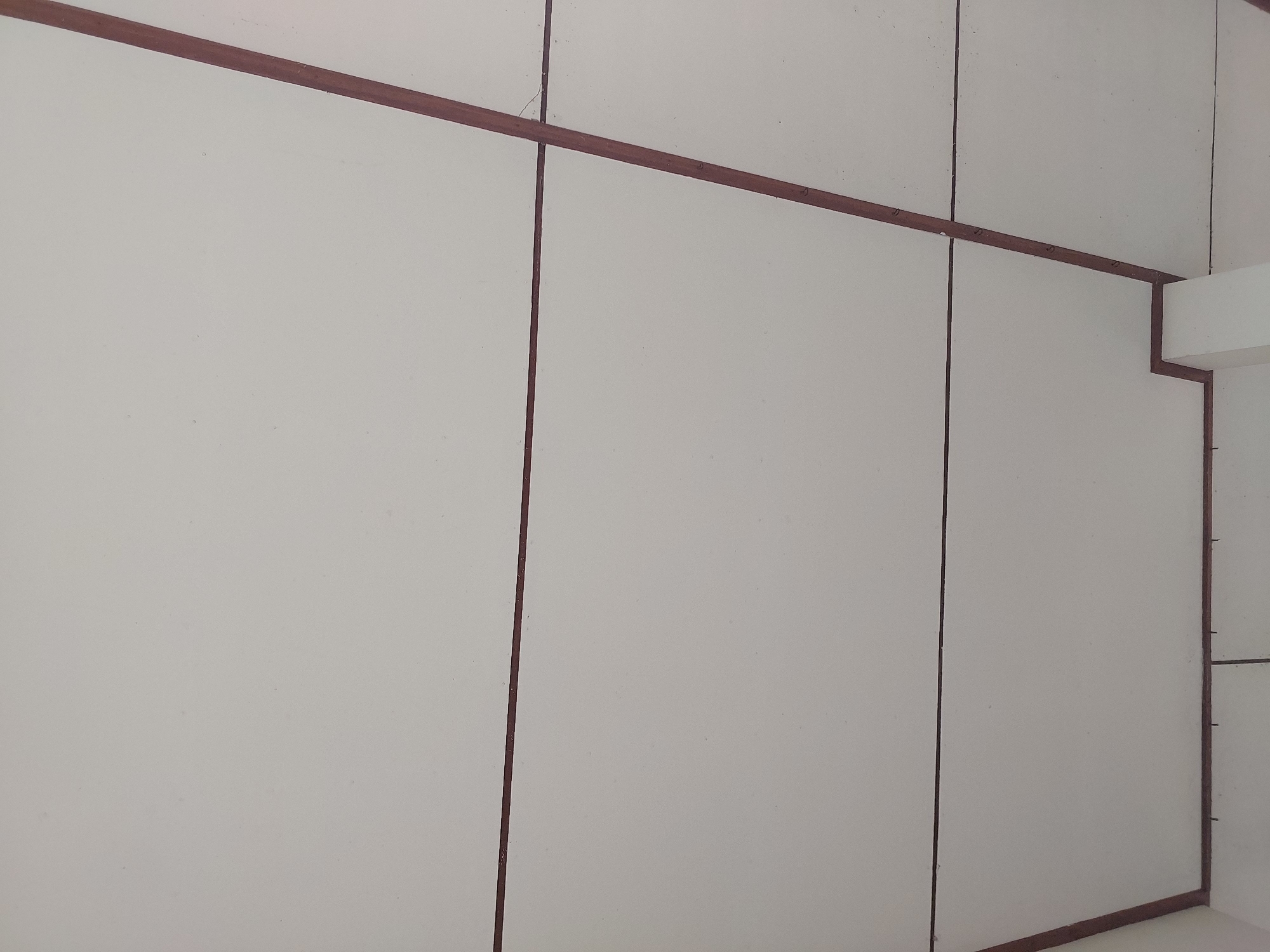 A low-angle, indoor, eye-level shot shows a portion of a white tiled ceiling with visible dark brown grout lines dividing the tiles. The ceiling appears to be suspended, as suggested by a white column visible in the lower right corner. The grout lines form a grid pattern, with one diagonal line prominent in the right half of the image. The lighting is even and diffuse, indicating an interior space with no direct light source visible. The overall scene is one of a simple, functional ceiling in what appears to be a building in Baan Mae Nam, Thailand.