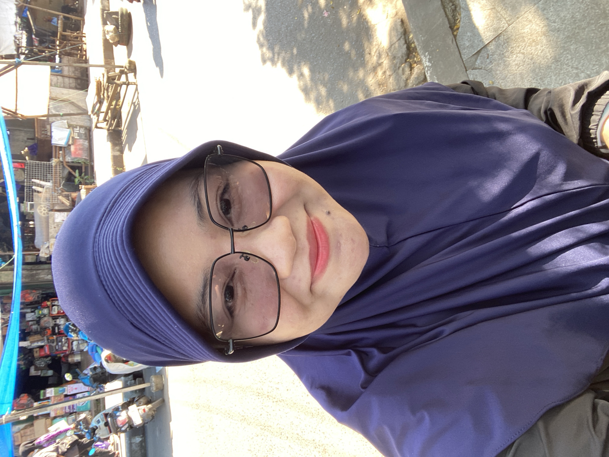 A woman wearing a dark blue hijab and glasses takes a selfie on a sunny day in Martapura, Indonesia. She is smiling and looking at the camera, with her face partially obscured by her hijab. The background shows a street scene with market stalls, motorcycles, and trees, suggesting a bustling urban environment. The woman appears to be standing on a sidewalk.