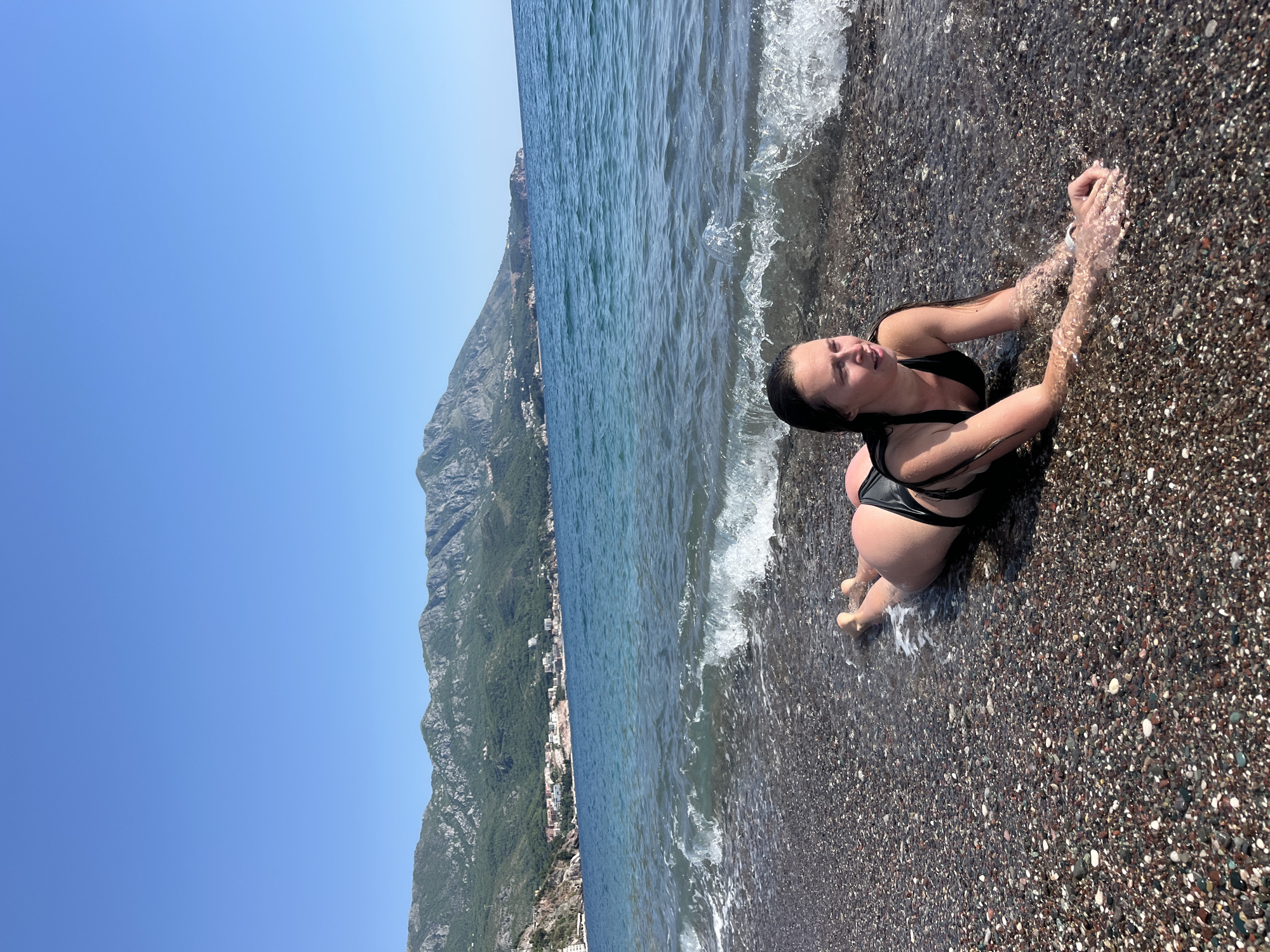 A woman in a black bikini lies prone on a pebble beach in Boreti, Montenegro. Waves wash over her hands and feet. The Adriatic Sea extends to the left, with a large mountain covered in greenery and housing small buildings along its base. The sky is clear blue.