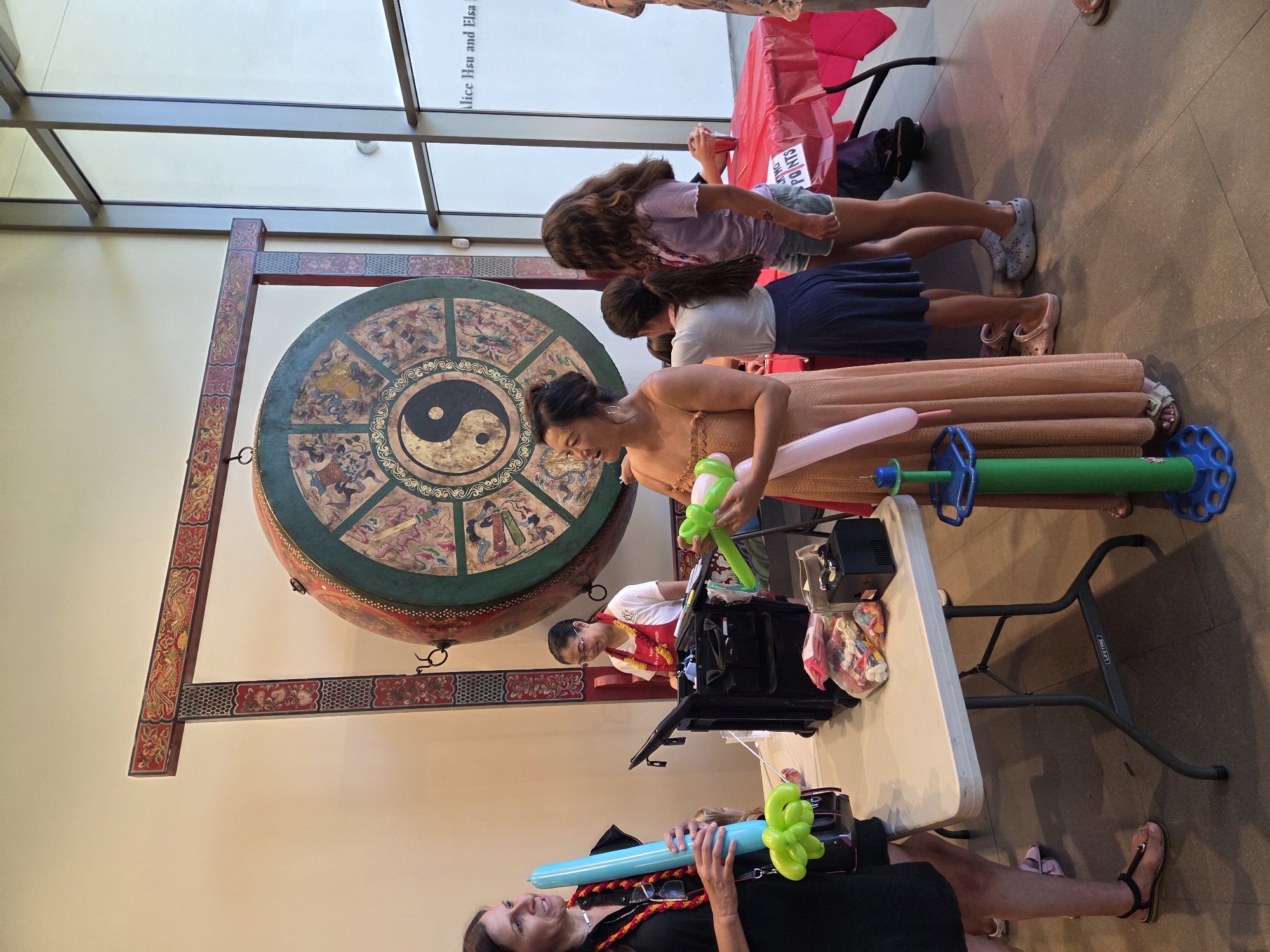 In Santa Ana, California, at an indoor event, several people are gathered. A woman on the left, wearing a black dress and a colorful necklace, holds a light blue balloon animal. Beside her, a child's legs are visible. In the center, a woman in an orange dress and another in a white shirt with a red apron are interacting near a black rolling cart filled with balloons. A large, ornate drum with a yin-yang symbol is displayed on the wall behind them. To the right, two young girls, one in a purple top and denim shorts, the other in a white top and blue skirt, stand near a table covered with a red tablecloth, where a sign reads "KING POINTS." Other objects in the scene include a balloon pump and a small black machine. The environment appears to be a public space, possibly a museum or community center, with large windows and tiled flooring.