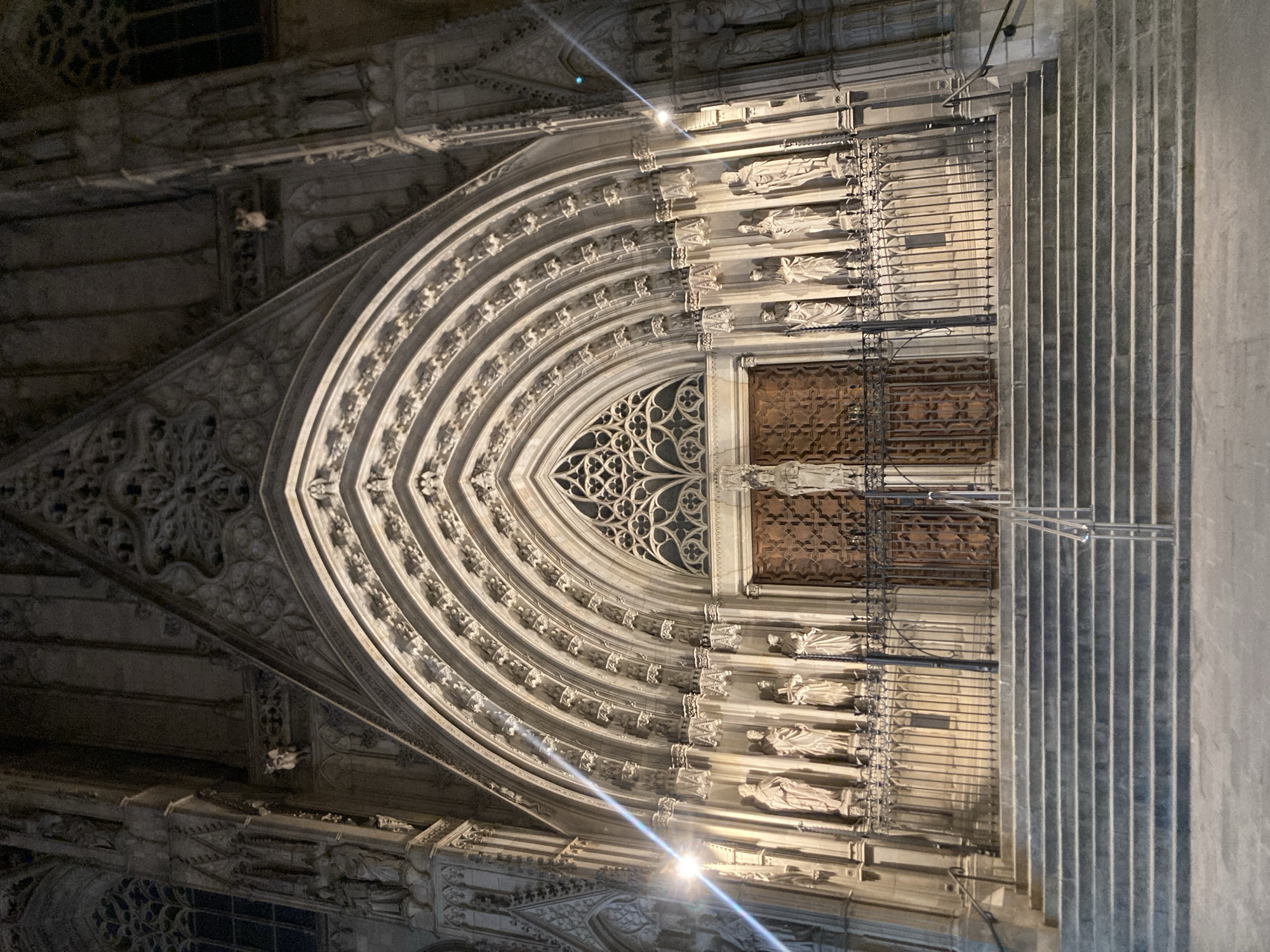 The image depicts the entrance of a grand Gothic cathedral, likely Barcelona Cathedral, in Barcelona, Spain, at night. The facade features an elaborate, multi-layered arched portal adorned with intricate carvings and numerous statues. A set of wide stone stairs leads up to the entrance, which is partially obscured by a decorative metal railing. Two large, ornate wooden doors form the main entrance, topped by a gothic rose window. The scene is illuminated by artificial lights, creating dramatic shadows and highlighting the architectural details of the cathedral. The ground in front of the stairs appears to be paved.