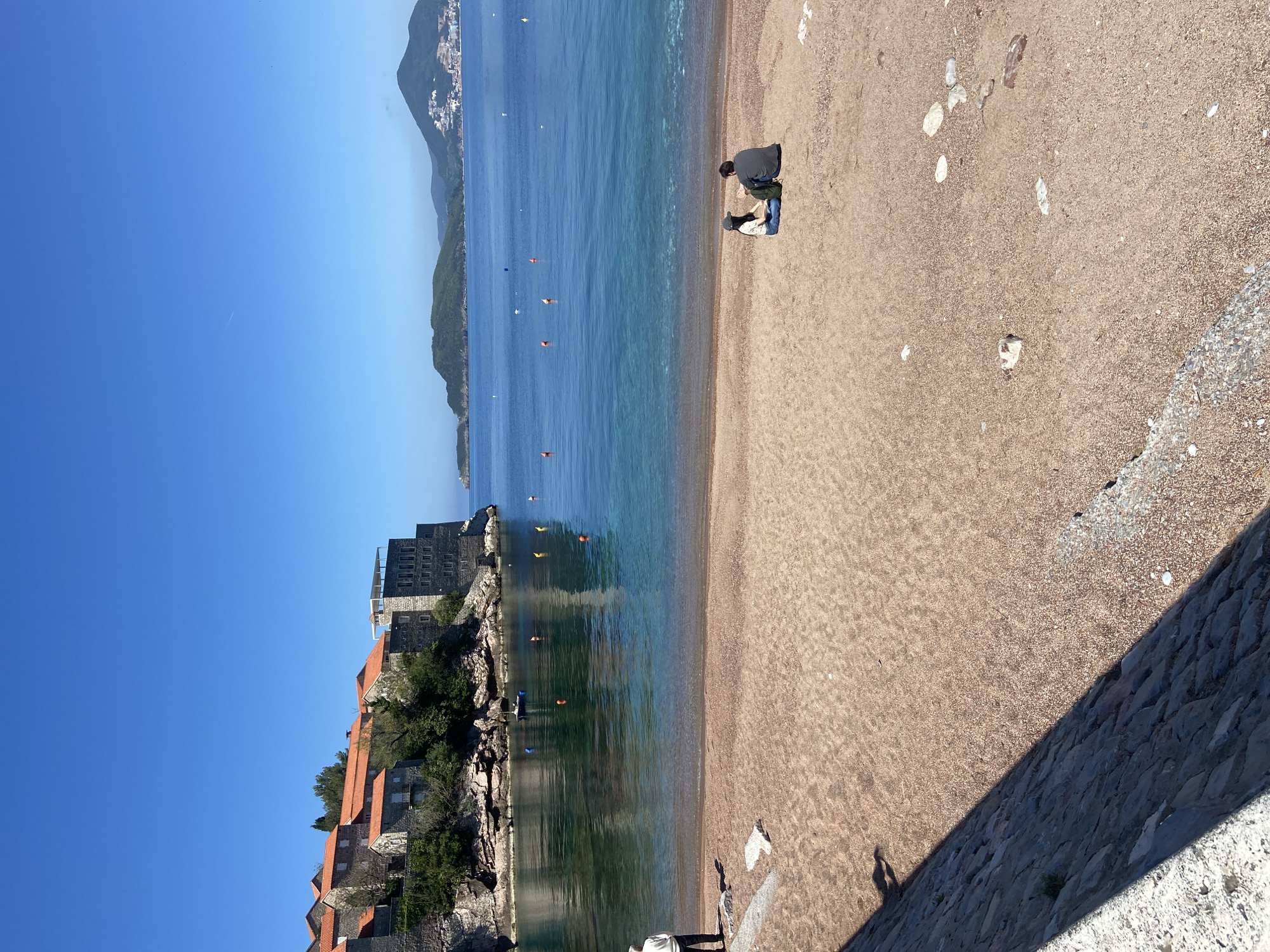 The image shows a beach scene in Sveti Stefan, Montenegro. The photograph is oriented with the beach on the right side and the water on the left. The beach is sandy, with a couple of people in the center right appearing to be looking at something on the sand. Scattered rocks and small pebbles are visible on the beach. The water is clear and blue with several orange buoys. The shoreline is rocky and lined with buildings featuring orange tile roofs and stone walls, with the taller building toward the back having several floors and what appear to be balconies. A mountainous green landscape is visible in the distance across the water. The sky is clear and blue. A stone wall can be seen in the lower right corner of the image.