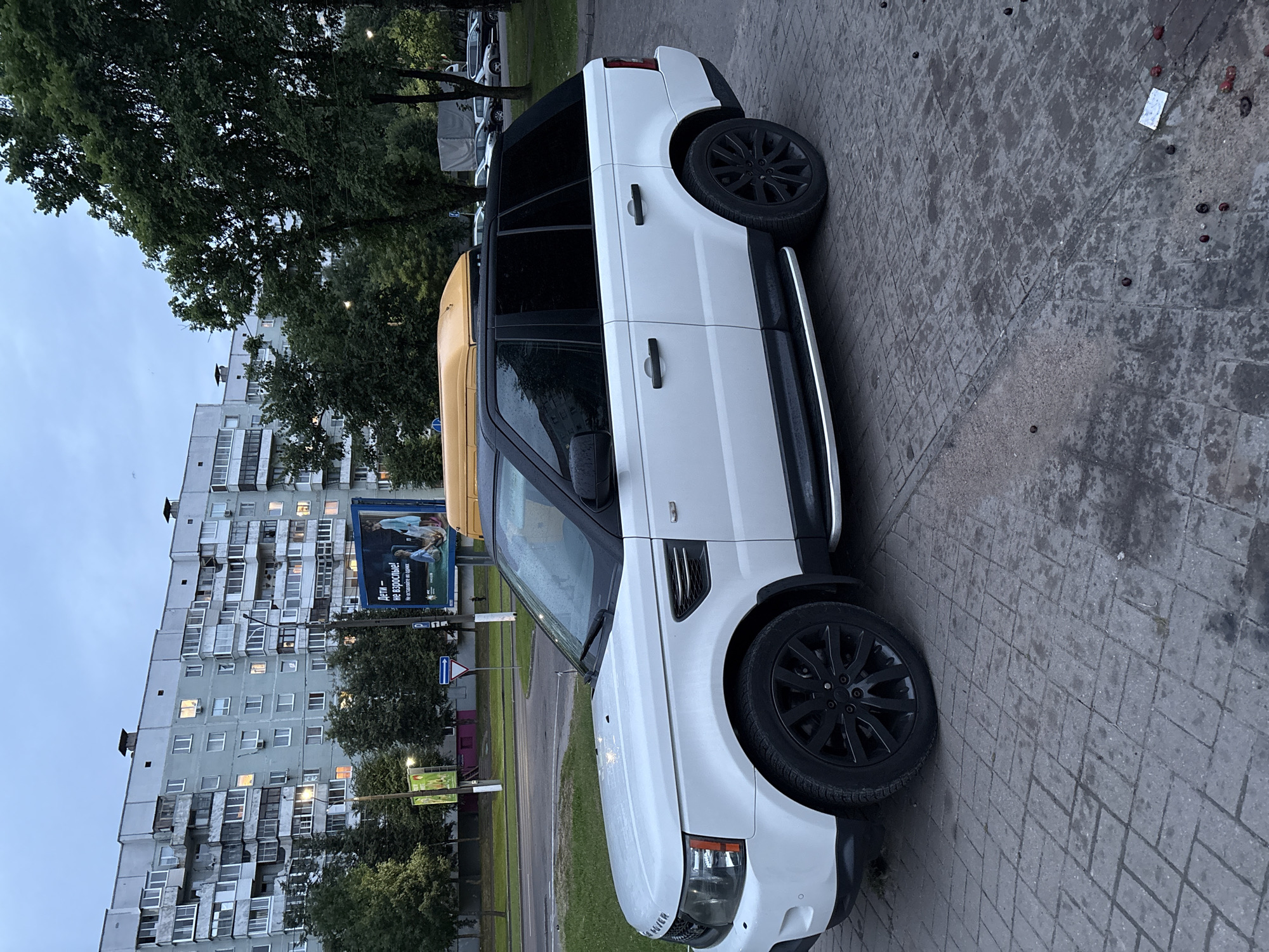 A white Range Rover Sport is parked on a wet, paved sidewalk in front of a tall, grey apartment building. The vehicle has blacked-out windows and black rims on its wheels. To the left of the Range Rover, a yellow trailer is partially visible. Beyond the trailer, trees and a blue and white billboard are seen. The billboard displays a picture of two people and text in Russian, with the words "Дети - не взрослые! Не становитесь в один ряд." The apartment building is multi-story with many windows, some of which are lit, suggesting it is either early morning or evening. The sky is overcast, indicating cloudy weather, and the wet pavement suggests recent rain. The scene appears to be a residential area in Vitsebsk, Belarus.