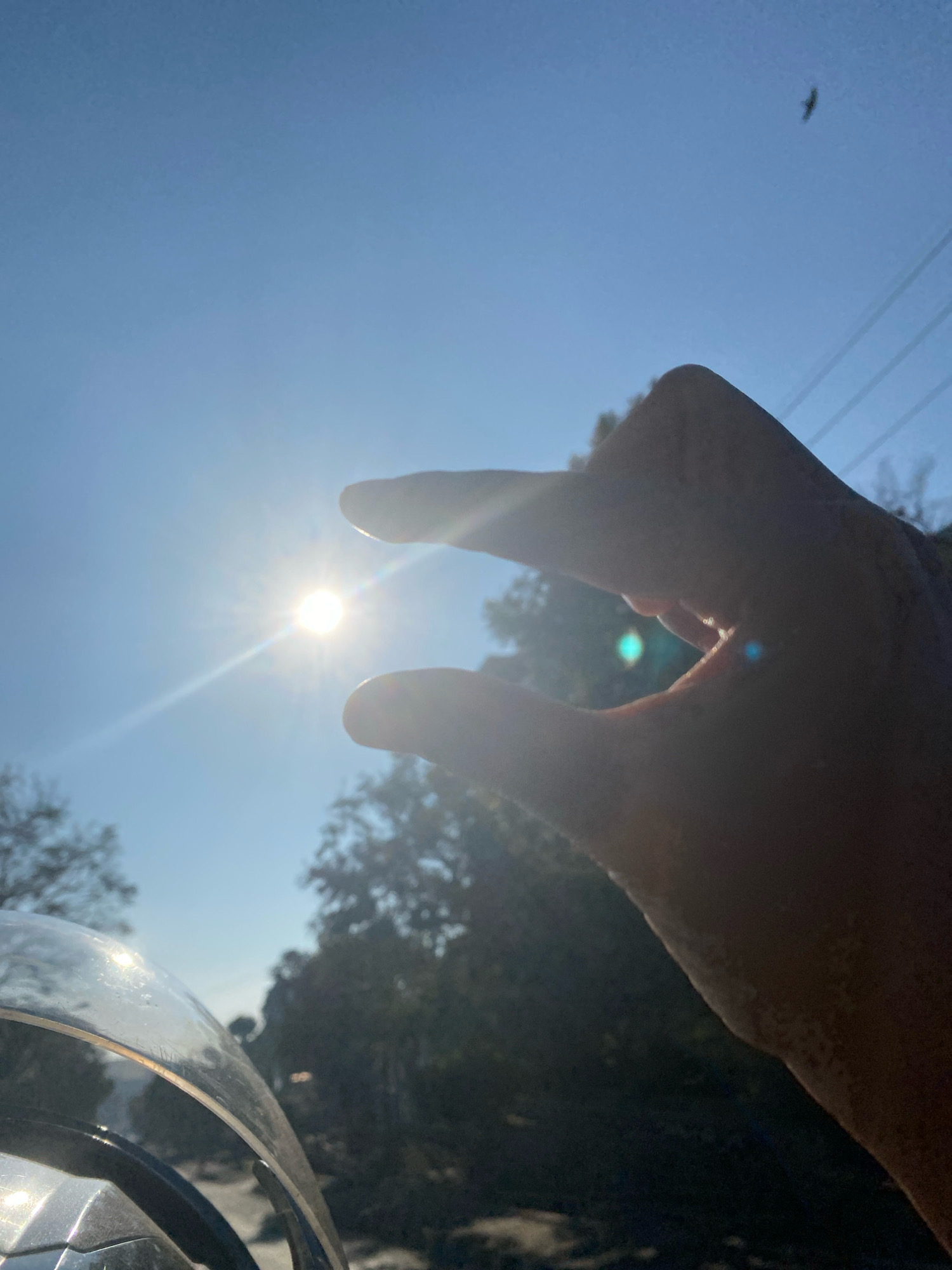 A close-up, low-angle shot captures a hand forming a pinching gesture with the thumb and index finger, obscuring a bright sun in the sky. The sun, a white-hot disc, is positioned in the upper left quadrant of the frame and emits a strong lens flare that streaks diagonally across the sky. The sky is a clear, pale blue. In the background, the silhouettes of trees and what appear to be utility lines are visible against the bright sky. A small, dark shape, possibly a bird, is visible in the upper right portion of the sky. In the foreground, a portion of a reflective surface, likely a motorcycle windshield, is visible at the bottom left, also reflecting the sunlight. The overall impression is a sunny day, possibly taken from a moving vehicle.