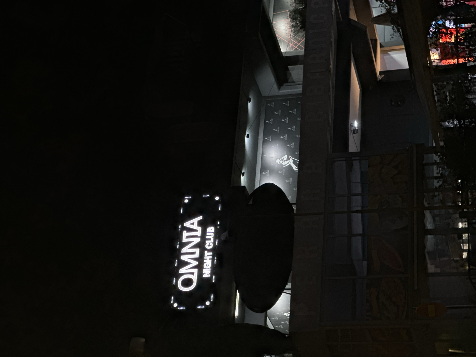 The image shows a nighttime view of the Omnia Night Club in Budva, Montenegro. The illuminated "OMNIA NIGHT CLUB" sign is prominently displayed. The architecture includes modern elements and a structure with the inscription "RIBARNICA". Other visual details are partially obscured by darkness. The presence of trees and reflections of lights from a nearby building or structure are seen. The environment suggests an urban setting at night.