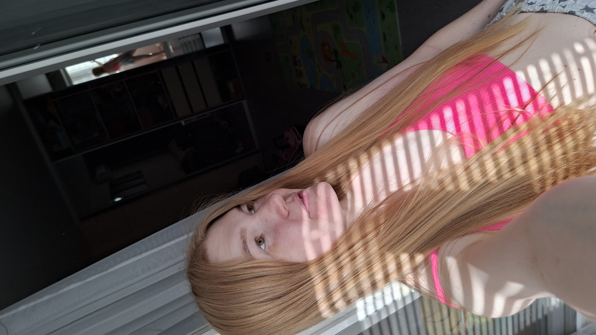 A woman with long red hair is lying down. She is wearing a pink top. The image appears to have been taken indoors near a window. In the background, there's a dark storage unit and a colorful child's play mat. The location is unknown.