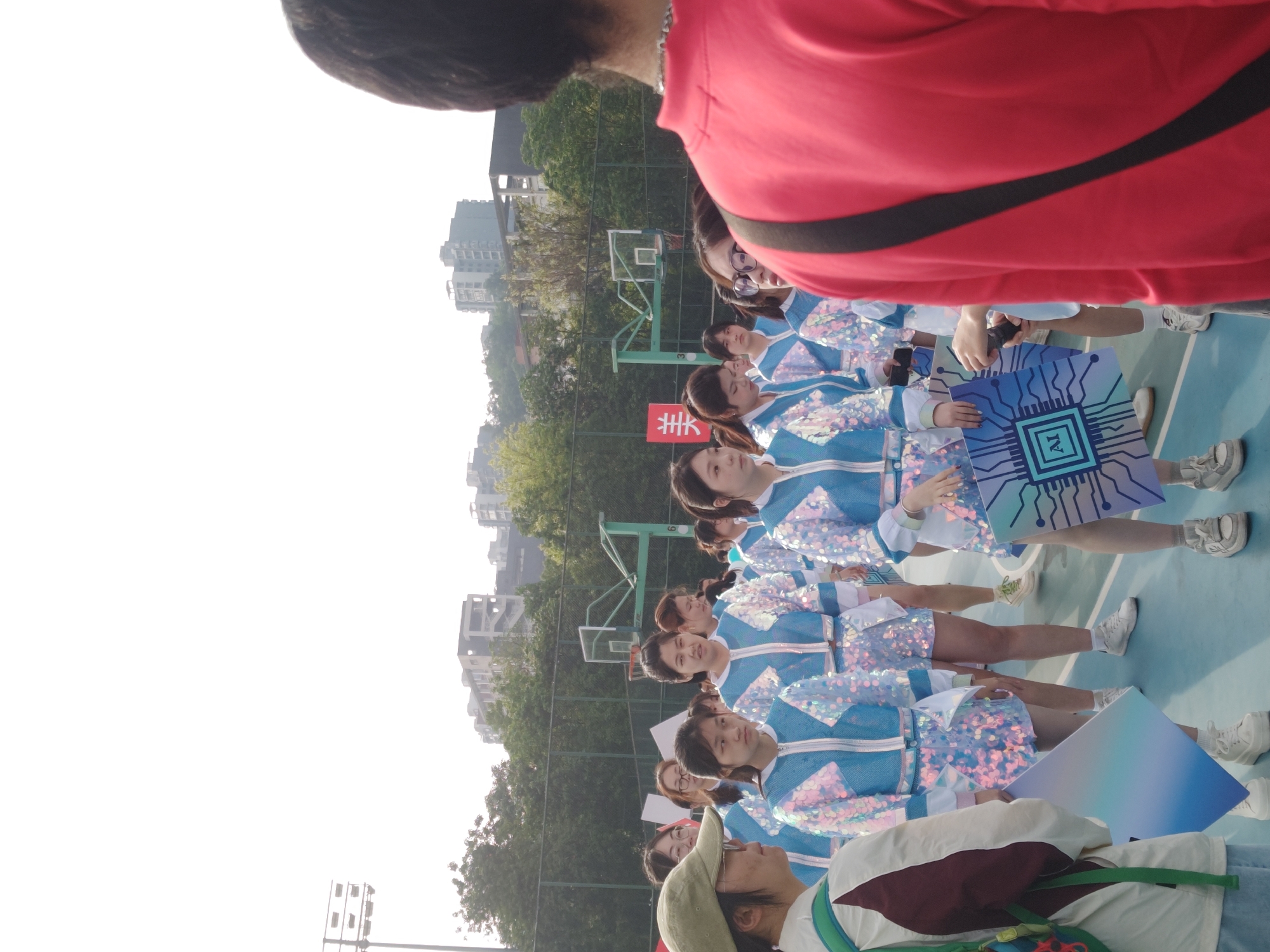 The image shows a group of young women standing in a line on an outdoor basketball court. They are wearing matching blue and white sequined outfits with white sleeves and are holding large, colorful signs that appear to depict computer chips with the letters "AI" on them. In the background, there are trees and buildings, suggesting an urban setting. A red banner with a Chinese character is visible. There is a person wearing a red shirt standing in front of them. The scene is taking place in the city of Chongqing High-tech Zone, China.