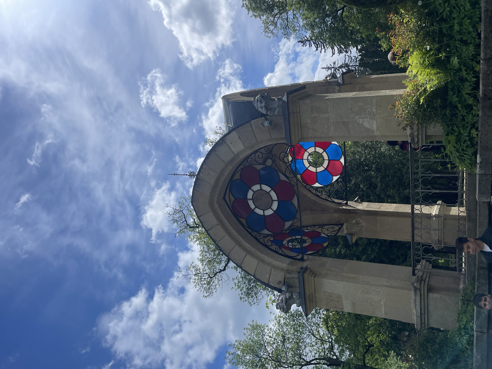 The image captures a unique architectural structure in Kislovodsk, Russia. This structure, potentially a gazebo or monument, is adorned with colorful circular stained glass windows, featuring a red and blue pattern. The building's exterior is made of light-colored stone, with decorative gargoyles on its upper corners.
The background reveals a clear blue sky with scattered white clouds, and the presence of green trees and shrubbery adds to the natural environment. There are hints of people in the scene, though they are partially obscured by the structure and foliage. The orientation of the image suggests the shot was taken at an upward angle, emphasizing the height of the architectural piece against the sky.
