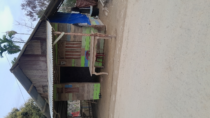 The image displays a weathered, single-story wooden building located along an unpaved road in Rumohrayeuk, Indonesia. The structure features a corrugated metal gable roof and an extended front awning. Its facade consists of horizontal wooden planks painted in faded green, blue, and natural wood tones. A dark, open doorway is centered, flanked by two window openings fitted with vertical wooden slats. An electrical meter box is mounted on the wall to the left of the door. A light-colored wooden table with ornate legs is positioned directly in front of the doorway, with a single red plastic chair placed behind it. On the right side of the building, a blue tarp covers a portion of the wall, and a dark bucket rests on the ground nearby. A rustic wooden post supports the right side of the front awning. Several pairs of sandals are visible on the dirt ground near the building's entrance. Lush green trees and several tall palm-like trees are visible in the background under a light sky. No individuals are present in the scene.