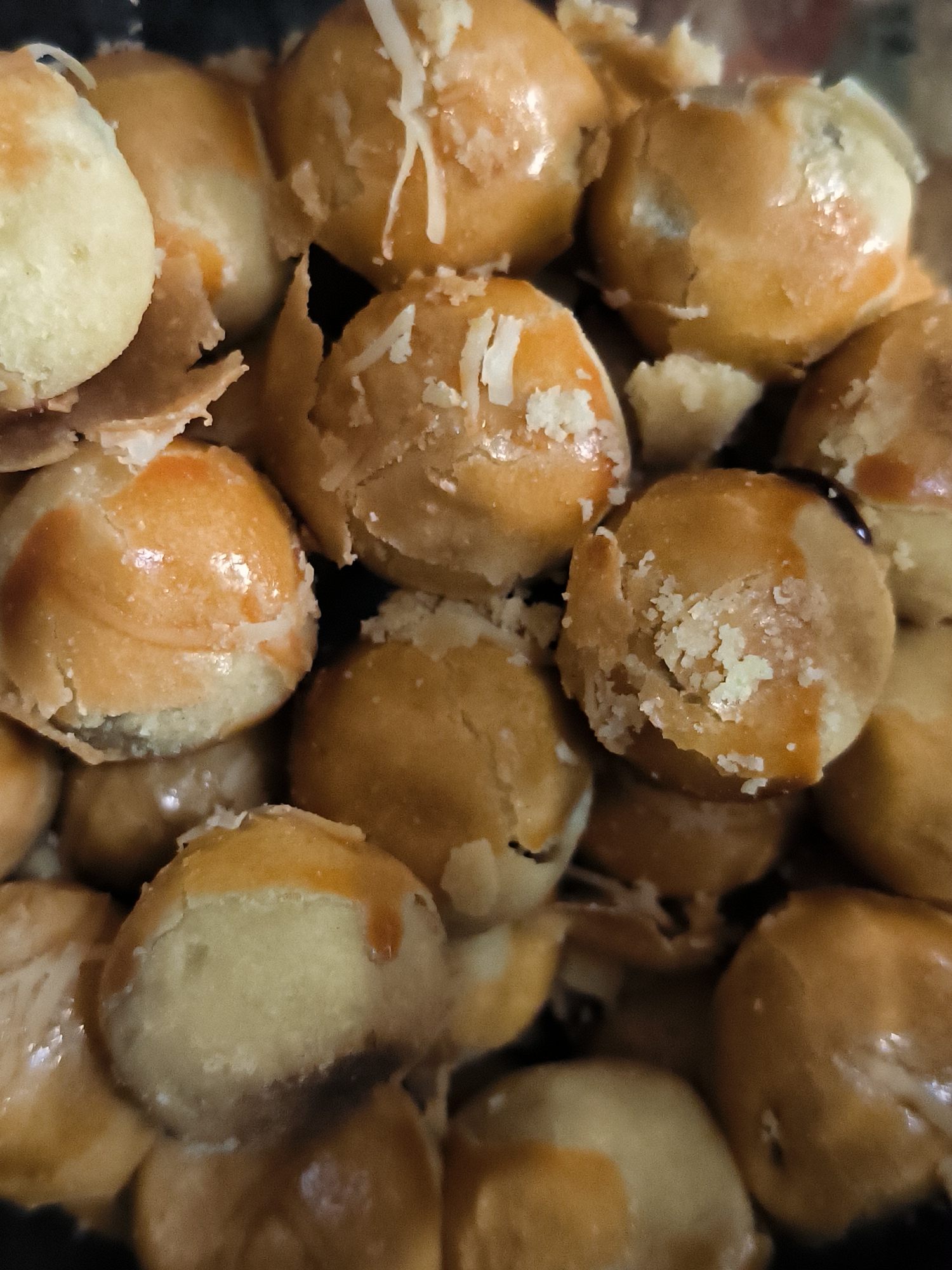A close-up vertical shot displays a dense arrangement of numerous small, round baked goods, resembling pastries or cookies. Each item, approximately 3-5 cm in diameter, features a light golden-brown crust, with visible cracks and irregularities on the surface. Many of the pastries are topped with a fine scattering of white, shredded material, possibly grated cheese or coconut, enhancing their texture. Some items exhibit areas of lighter, off-white dough, suggesting either exposed interiors or a two-toned baking effect, along with a glossy finish that indicates a glaze. The individual pastries are tightly packed, with some appearing partially broken or crumbled, revealing lighter interiors. The background is dark and indistinct, suggesting a shallow depth of field. This scene is observed in Lhokseumawe, Indonesia.