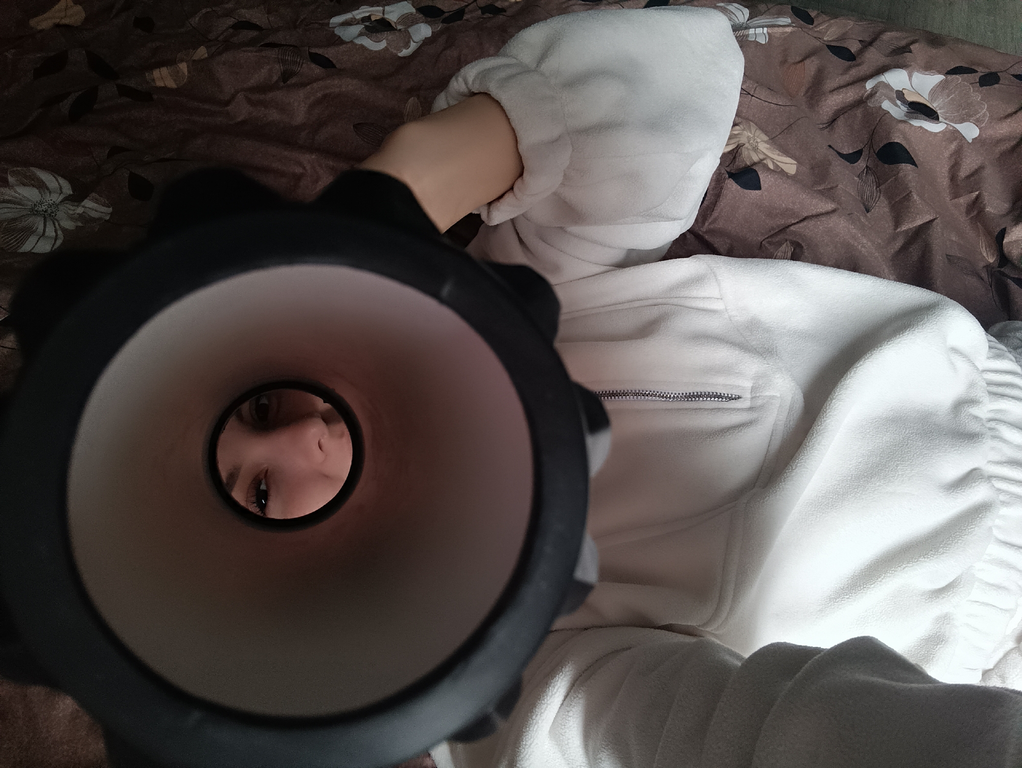 An overhead shot captures a person lying down, looking into the open end of a cylindrical object. The main subject is a person, whose face – specifically the eyes, nose, and forehead – is visible reflected or directly viewed through the narrow opening of a dark-colored, possibly black, cylinder. The person's arm, with light skin tone, extends from a white, plush fleece garment towards the cylinder. The upper body of the person is clad in a white fleece jacket or top, which features a silver zipper on the front, partially visible.
The setting appears to be indoors, likely a bedroom, as the person is lying on a bed covered with a patterned sheet or blanket. The bedsheet has a warm brown background adorned with a pattern of white, light brown, and dark brown stylized flowers and leaves.
The activity depicted is the person looking into or through the cylindrical object, creating a unique perspective where their face is framed within the object's interior. The overall impression is one of curiosity or engagement with the object. No text is visible in the image.