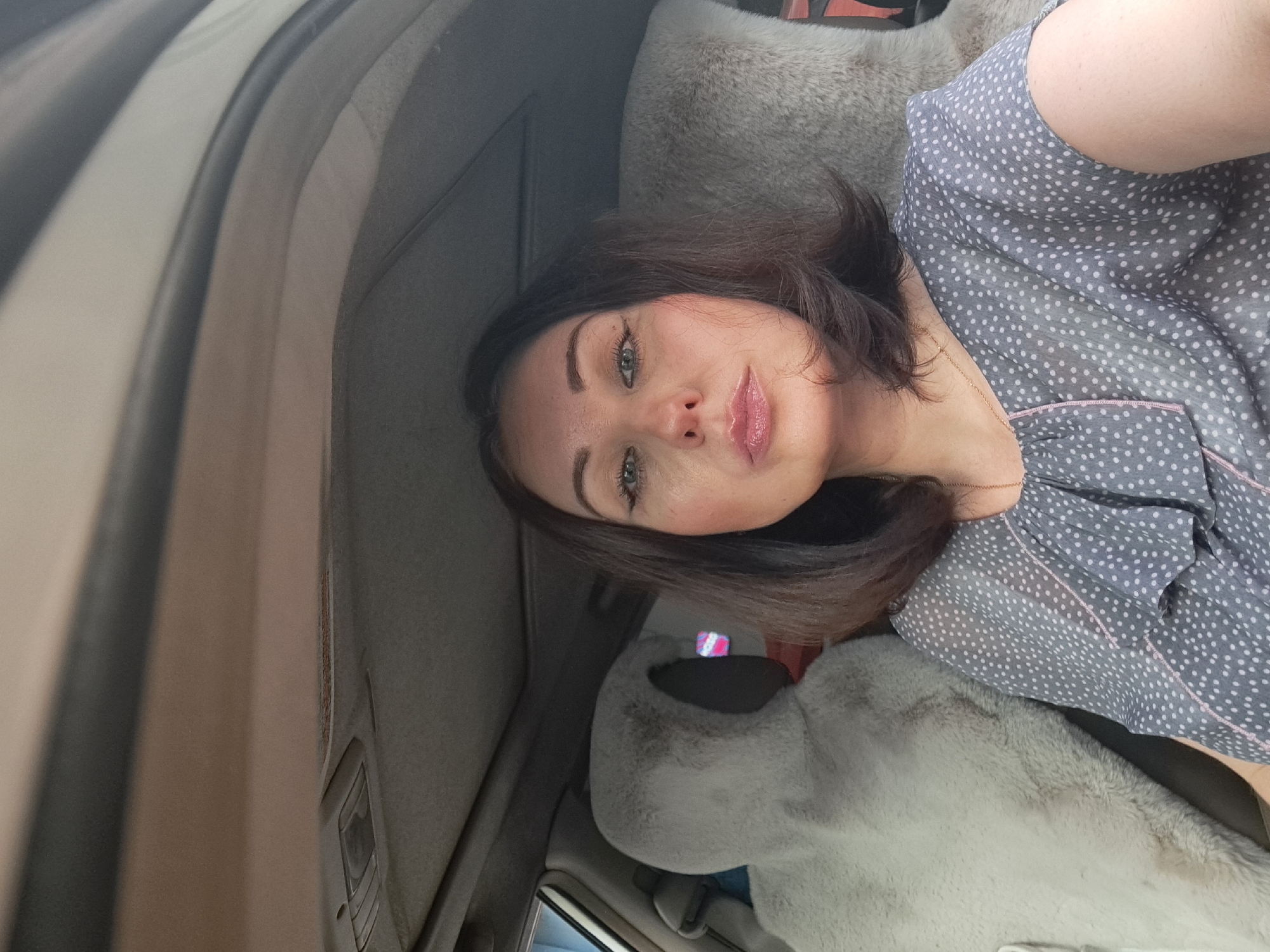 A woman is sitting in the driver's seat of a car, looking at the camera. She has dark brown hair, blue eyes, and is wearing a grey polka dot blouse. The car's interior is visible, including a fluffy grey sheepskin seat cover on the passenger seat. The image appears to have been taken in Irkutsk, Russia, as indicated by the context provided.