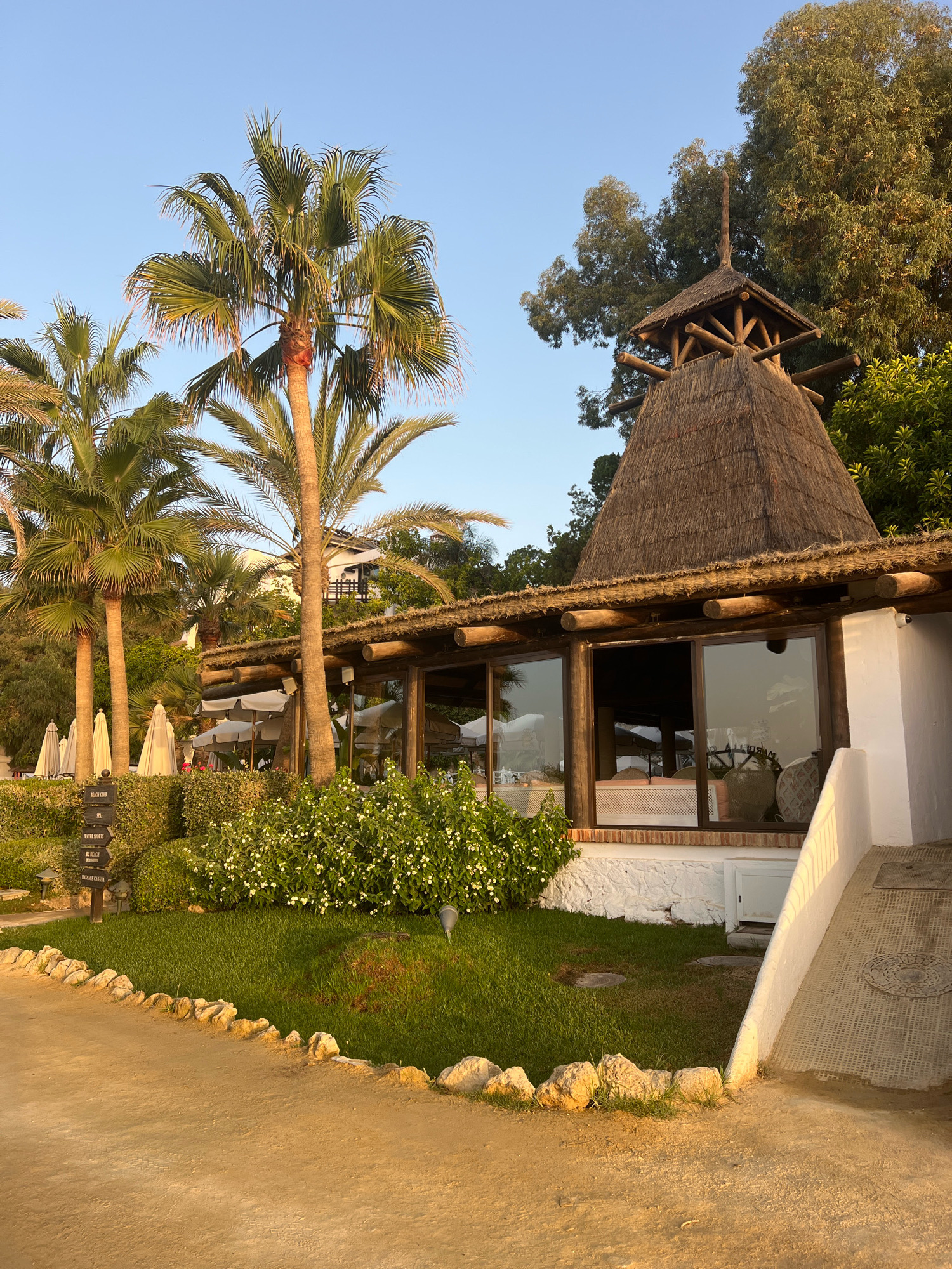 The image shows the exterior of a beachfront restaurant in Marbella, Spain, during what appears to be late afternoon or early evening. The restaurant has a rustic design with a thatched roof structure and large windows that look out onto a garden and palm trees. A pathway, made of light brown dirt, leads to the restaurant. To the left, a signpost indicates directions to "BEACH CLUB", "SPA", "WATER SPORTS", "BIG BEACH", and "MASSAGE CABANA". Several white umbrellas are visible behind a low hedge and in front of the building. The sky is a clear blue, suggesting good weather. The overall impression is one of a relaxed, upscale resort setting.