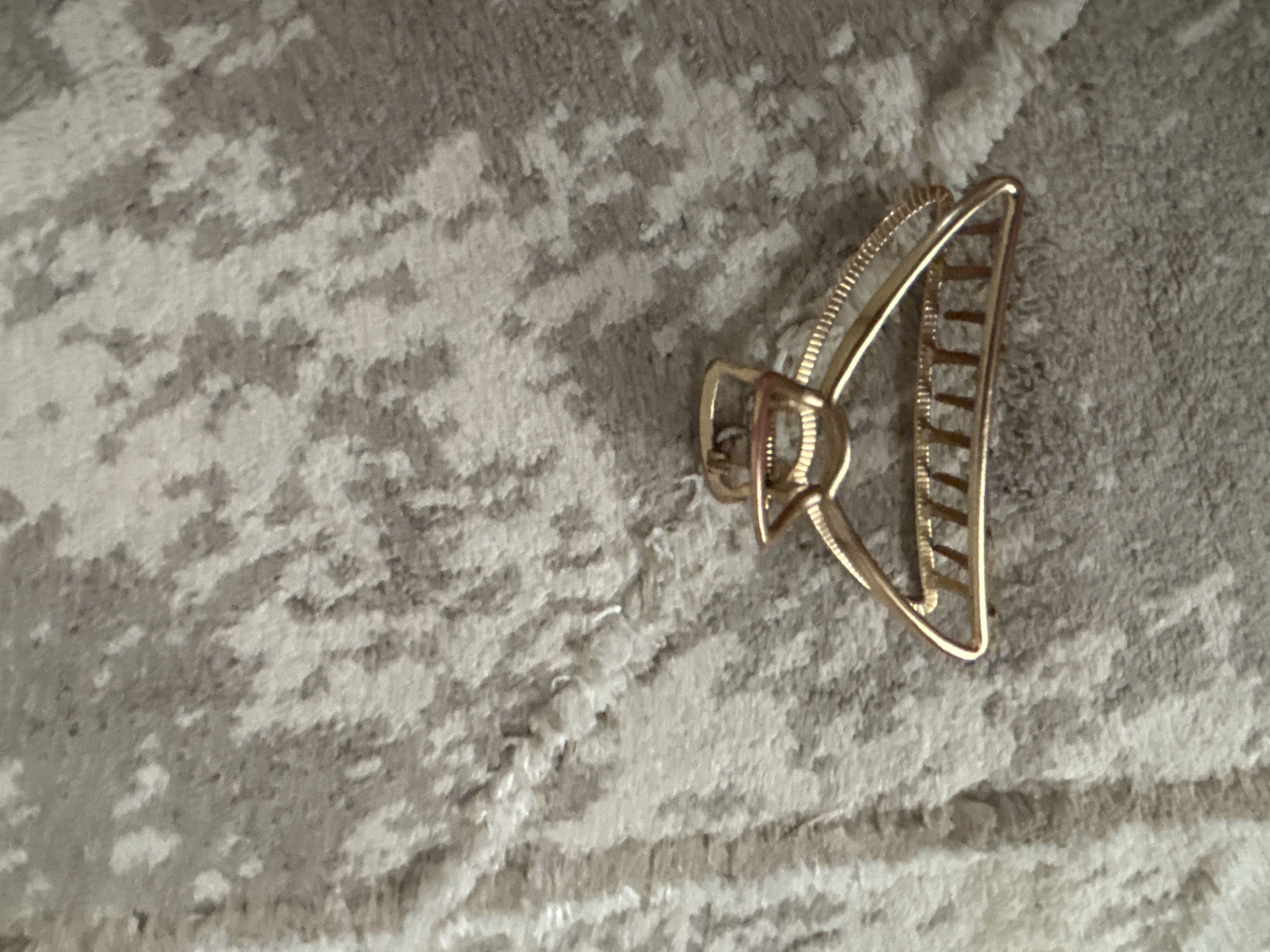 A close-up, overhead shot shows a gold-colored metal hair clip resting on a textured, light-colored rug. The rug has a pattern of lighter and darker gray splotches. The hair clip is open and positioned on its side, with its "teeth" facing upwards. The clip is made of shiny metal and appears to be a standard claw clip design. The image is taken in a way that the background fills most of the frame, with the hair clip as the central subject. The location in Kyzylorda, Kazakhstan is not discernible from the image itself, as there are no contextual clues.
