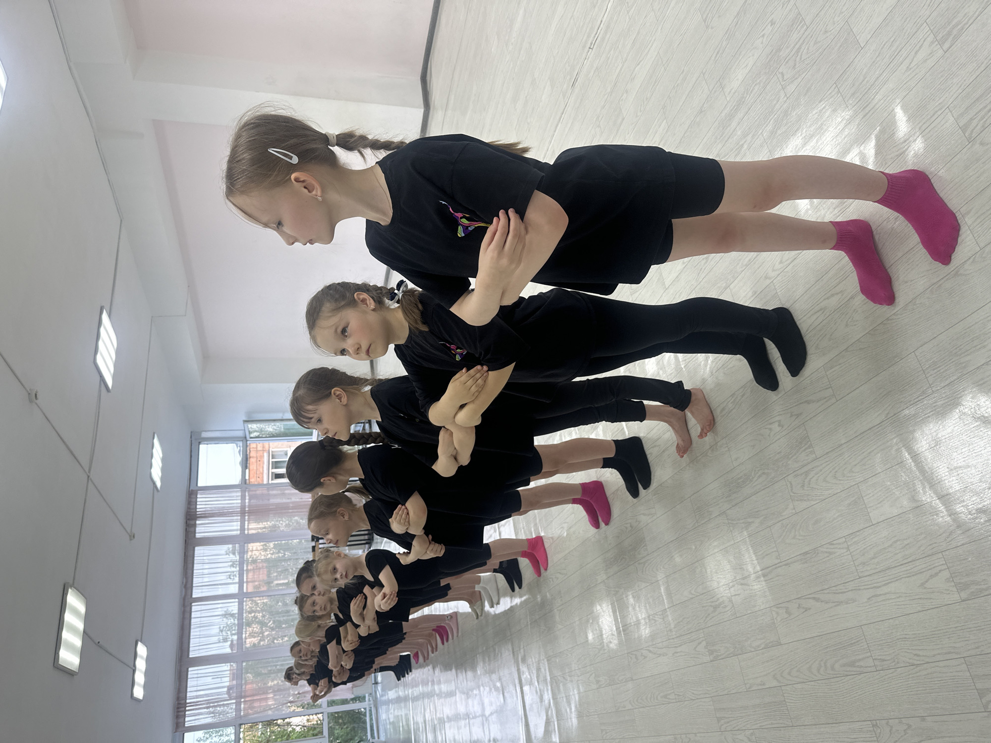 A horizontal, angled shot captures a group of approximately 12 young girls, lined up, in what appears to be a dance or exercise studio in Votkinsk, Russia. The girls are arranged in a row, each facing the same direction, with their bodies oriented towards the right side of the frame. All are wearing black attire consisting of a t-shirt and leggings. They are doing an exercise with their hands folded across their chest while in a plank-like position. Their choice of socks varies in shades of pink, black, and white.
The studio environment consists of light-colored, faux-wood-grain flooring, with a large window in the background to the left, partially obscured by building facades and greenery outside. A row of rectangular lights is suspended from the white ceiling, parallel to the line of girls. The overall lighting in the room is bright, creating a well-lit space.