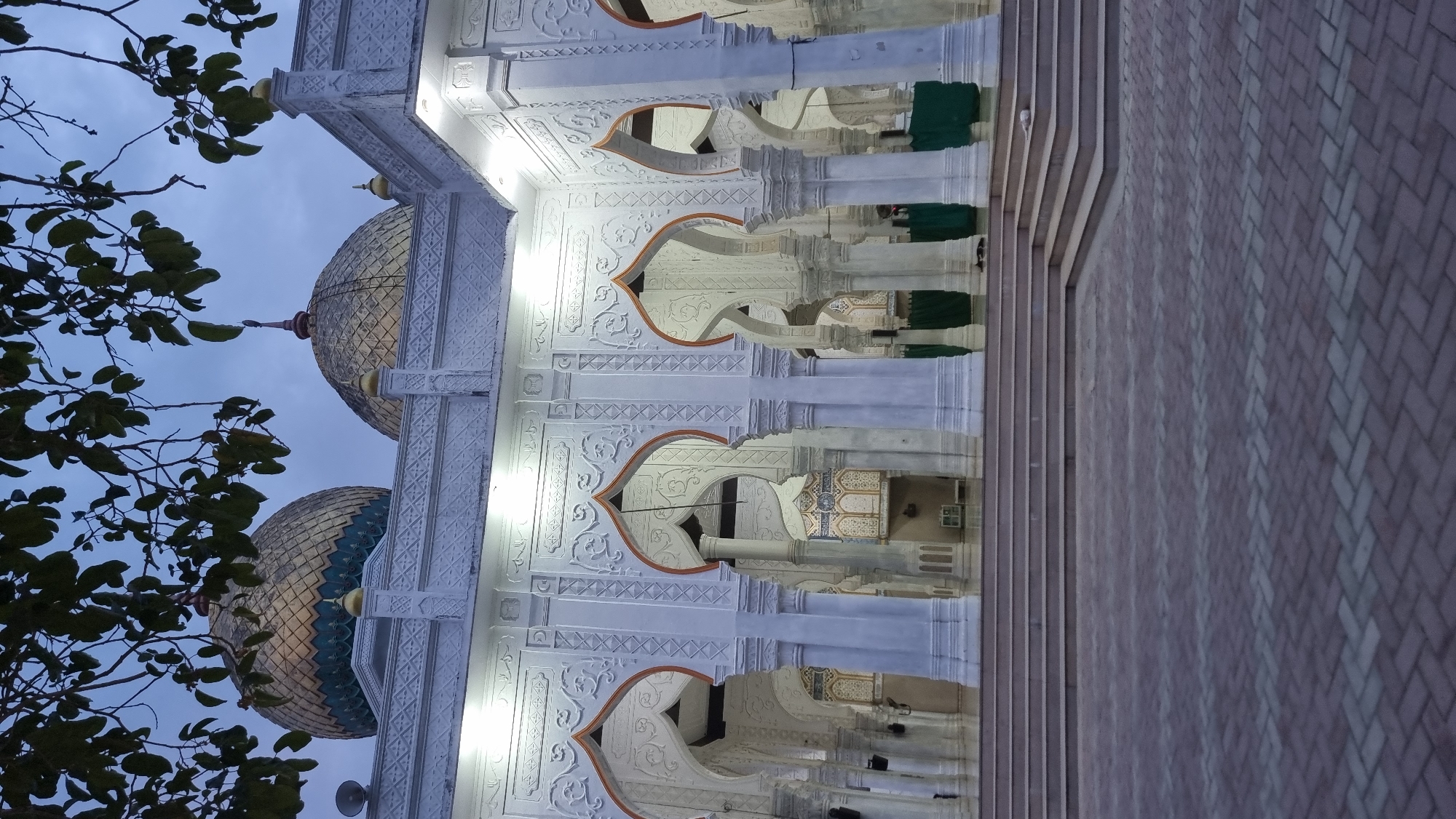 The image shows an exterior view of a mosque in Meunasahlhok, Indonesia, at dusk. The mosque features white walls with intricate relief carvings and several arched entrances supported by white pillars. Two prominent golden domes with teal accents are visible on the roof, partially obscured by dark green tree branches and leaves in the foreground. The ground in front of the mosque is paved with interlocking bricks arranged in a herringbone pattern. A set of wide stairs leads up to the mosque's entrance. The lighting suggests it is either early morning or late evening, with artificial lights illuminating the mosque's facade. No people are clearly visible in the scene.