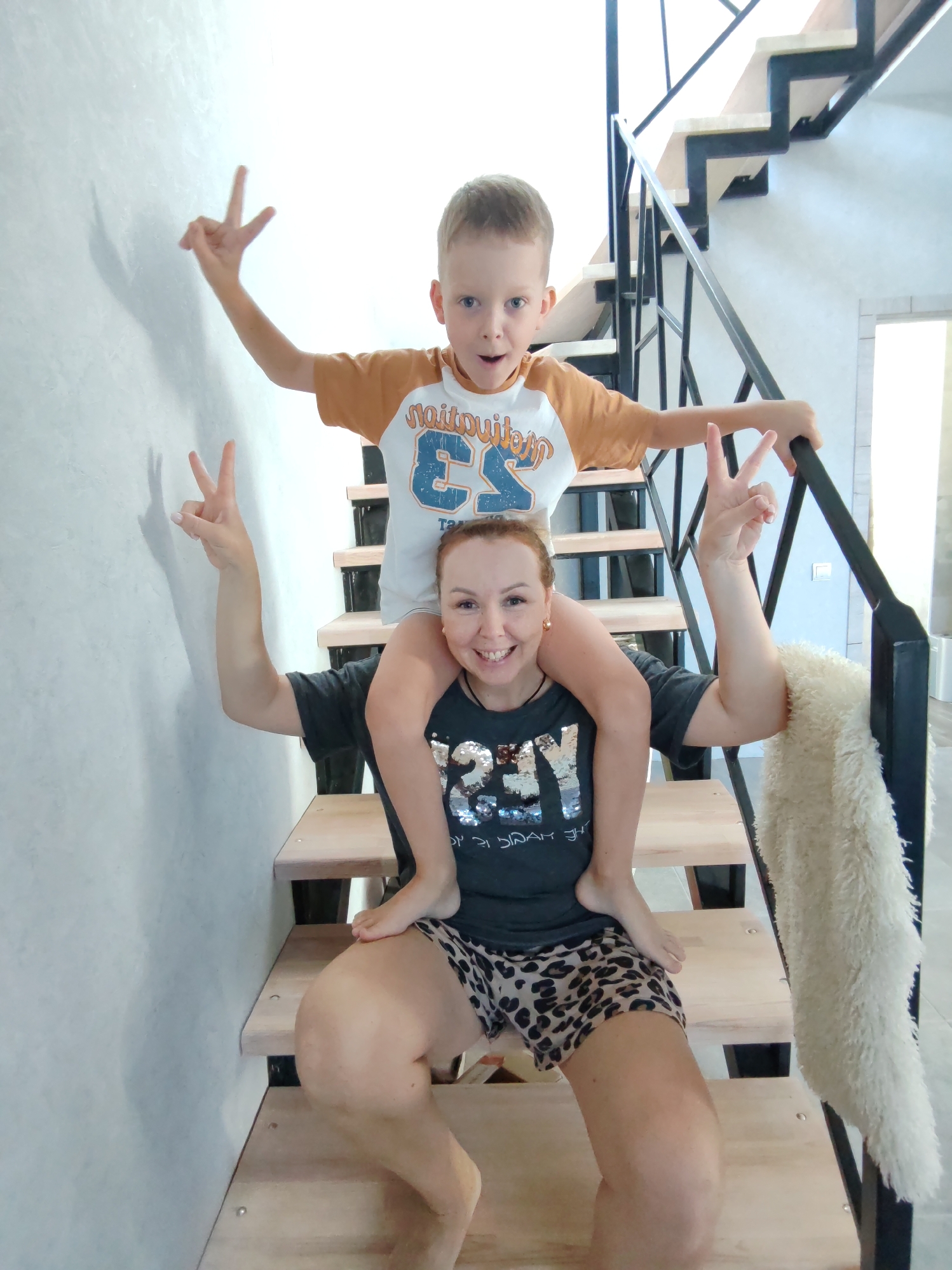 A boy is sitting on a woman's shoulders on a wooden staircase. The boy is wearing a brown and white t-shirt, and the woman is wearing a black t-shirt and leopard print shorts. Both are making a peace sign with their fingers. A fuzzy white blanket is draped over the railing of the staircase. The scene appears to be indoors, possibly in a residential setting. There is no indication of the city of Chaikovsky, Russia, in the image.