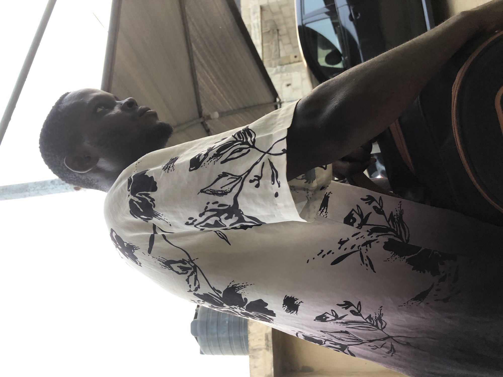 A low-angle shot depicts a dark-skinned man with short hair and a beard, wearing a light-colored, patterned shirt. He is looking upwards, with his head tilted back. Part of a car is visible in the background, along with a slanted, metallic structure. A backpack is partially visible on the right. The location appears to be outdoors in an urban environment.