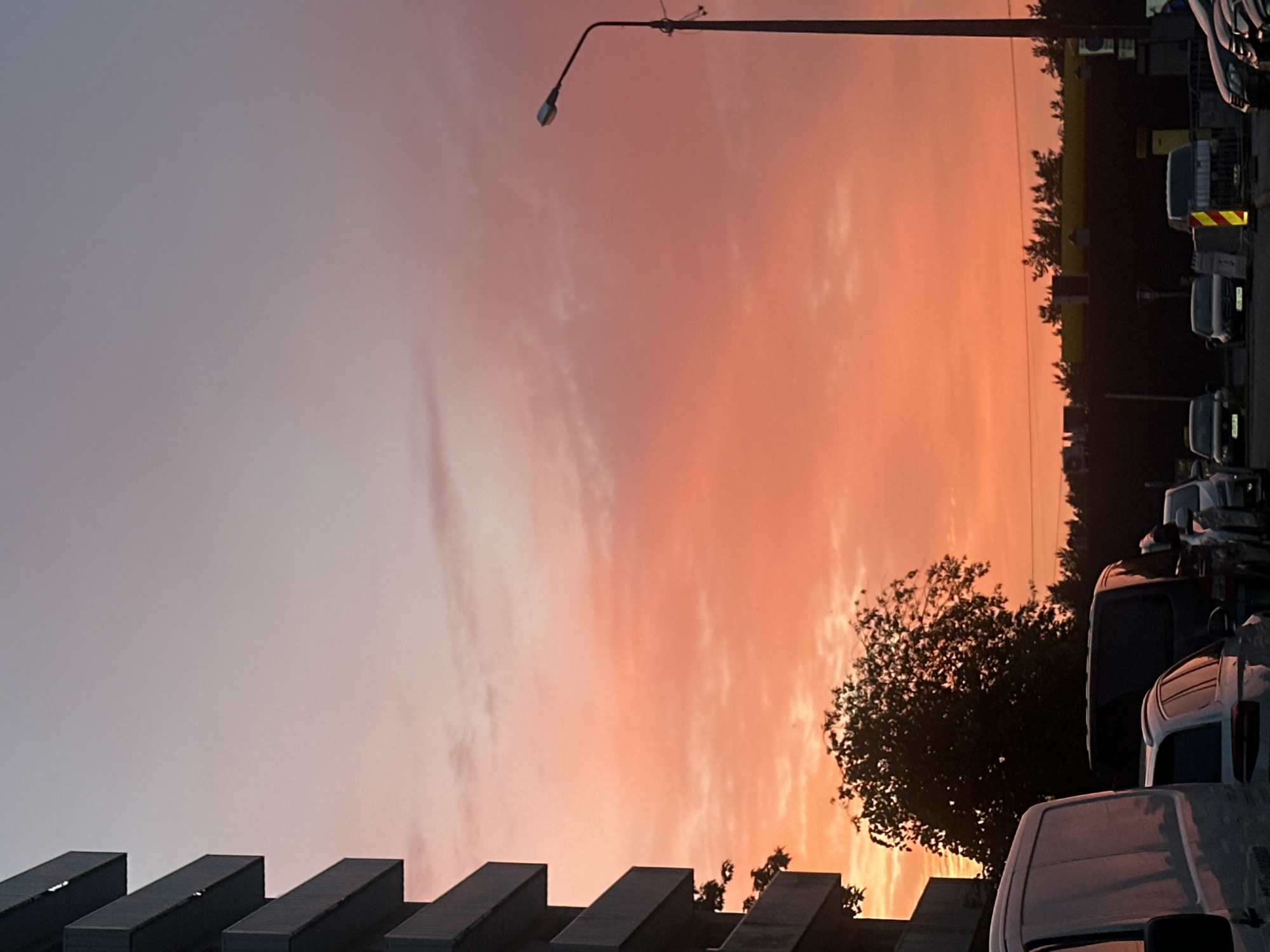 The image captures a sunset over a parking lot in Tallinn, Estonia. The sky is ablaze with hues of pink and orange, with a gradient fading to a pale blue at the top. On the left, a portion of a concrete building with multiple balconies is visible, partially obscuring the sky. In the foreground, several vehicles are parked, including vans and cars. A street lamp stands tall on the right side of the image, its light fixture illuminated against the colorful sky. A dense cluster of trees is silhouetted against the sunset in the middle ground, with more trees and buildings forming a dark line along the horizon. The scene appears to be a quiet evening in a city setting.