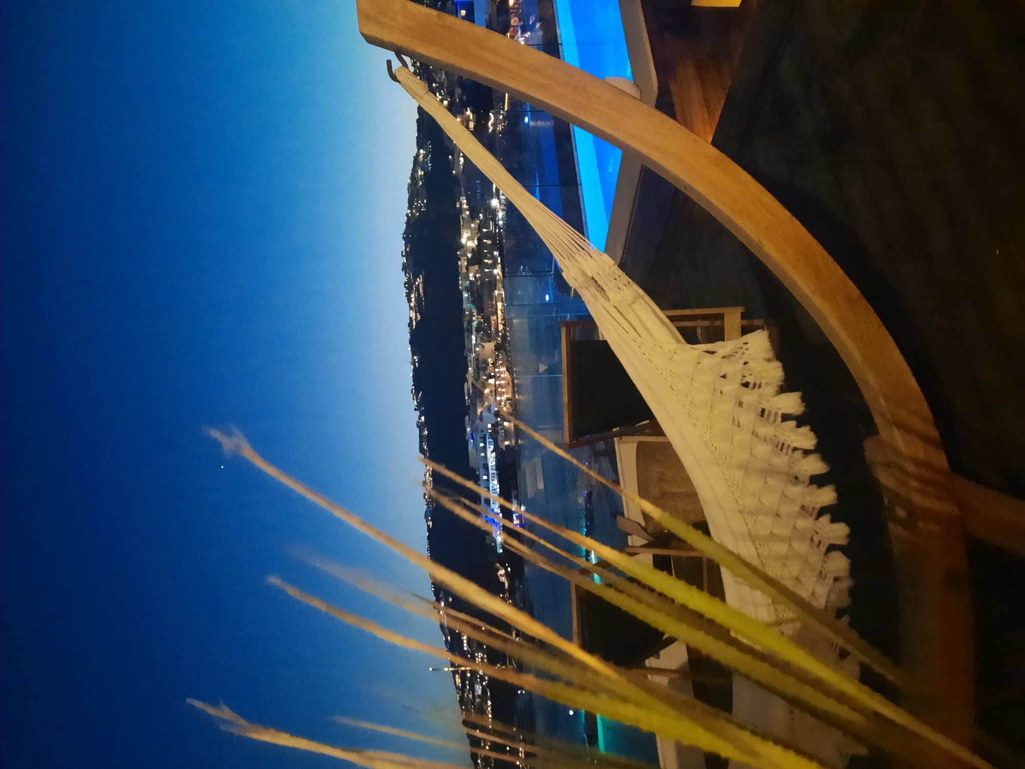 This is a nighttime photograph taken from a terrace overlooking the sea in Platys Gialos, Greece. In the foreground, a white hammock is suspended from a wooden frame. To the left, tall, thin blades of grass are blurred, suggesting they are close to the camera and swaying in the breeze. Behind the hammock, two dark lounge chairs are visible. The middle ground features the sea, with a body of water reflecting lights from a distant hillside town. The town is illuminated with numerous lights, indicating it is populated. In the background, a hill rises, covered with buildings and lights, suggesting a town or village. The sky is a deep blue, transitioning to a darker hue at the top, with a single bright star or planet visible. A bright blue swimming pool is situated to the right of the hammock, partially visible and illuminated from within. The overall scene conveys a sense of tranquility and relaxation at dusk or early night.