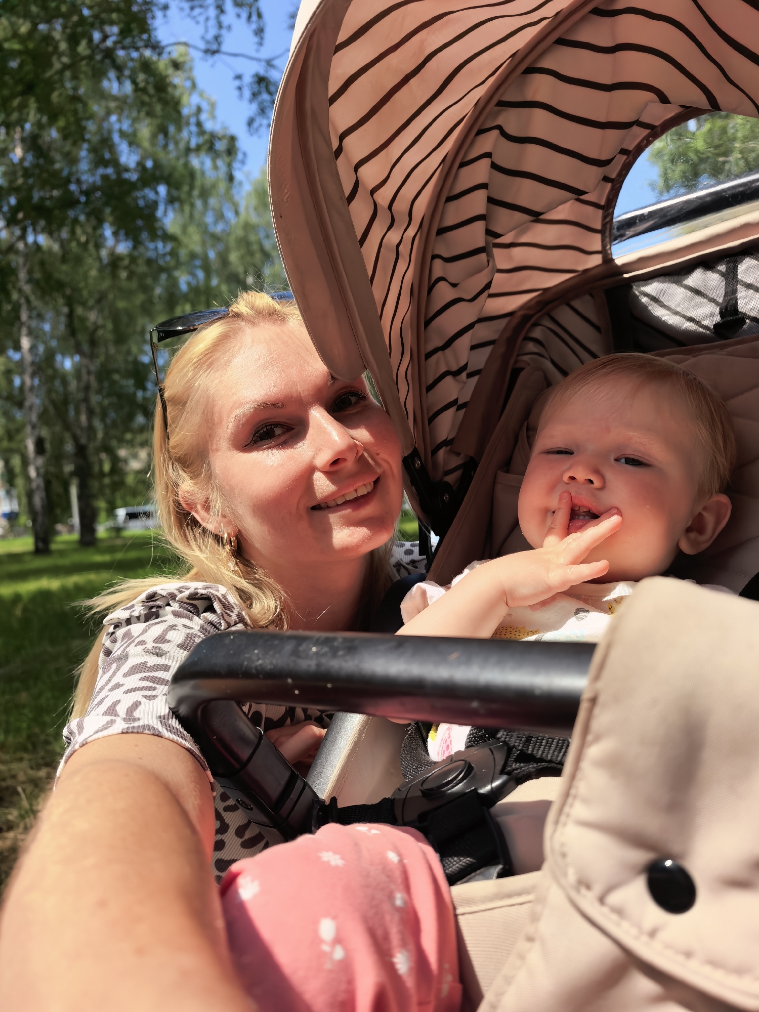 A woman and a baby are in a stroller outside. The woman is blonde and smiling at the camera. She is wearing a patterned shirt and has sunglasses on her head. The baby is in the stroller, facing the camera. The baby has its fingers in its mouth and is looking at the camera. The stroller is open and the baby is in a tan-colored seat. The background is a park with trees and a blue sky. The image appears to have been taken on a sunny day. The specific location is not identifiable.