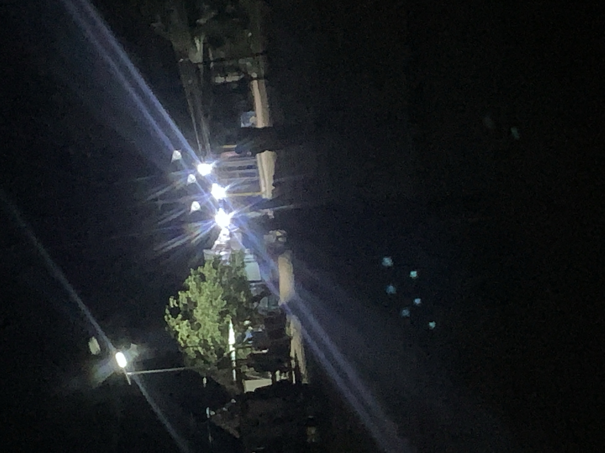 The image depicts a nighttime scene in what appears to be a public area, possibly a street or marketplace, in Jalingo, Nigeria. Several streetlights illuminate the scene, casting beams of light that create lens flares and streaks across the image. In the background, there are indistinct figures of people, some standing and others possibly moving. There are also structures visible, including what looks like a large tree with green foliage and some covered stalls or temporary shelters. The ground is dark, suggesting it's paved or dirt. In the foreground, there are several small, scattered points of light, some appearing to be greenish or bluish, which could be reflections or distant light sources. The overall impression is one of a moderately busy area at night, with artificial lighting being the primary source of illumination.