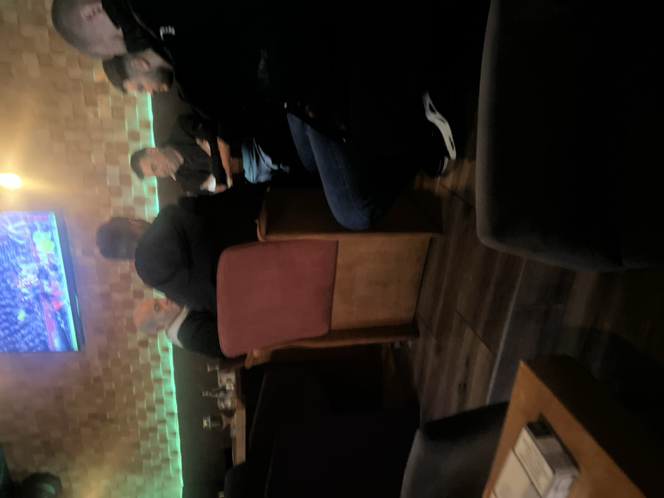 A group of people is gathered indoors, possibly in a bar, watching a television screen that displays a vibrant scene with green and blue hues. The room has a tiled wall illuminated with green light. A man in a suit sits in a maroon chair. A wooden table is visible on the floor. Dark sofas and armchairs suggest a comfortable, lounge-like atmosphere. Some people are standing, with one individual wearing blue jeans and white sneakers.