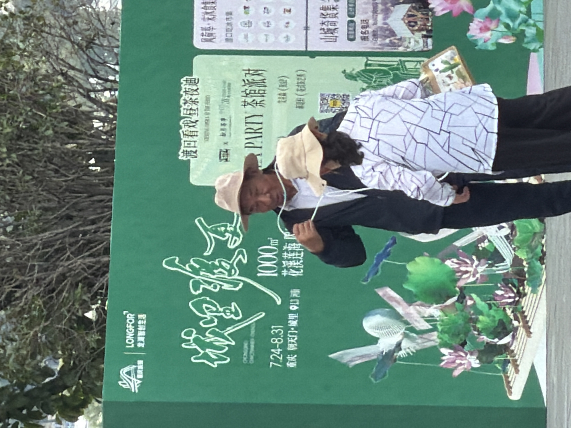 The image captures an outdoor scene with a large green banner serving as a backdrop. Two individuals, an older man and a woman, stand in front of the banner, appearing to be engaged in some activity involving a rope. The man, wearing a light-colored hat and dark jacket, has the rope draped over his hands and is looking towards the woman. The woman, also wearing a light-colored hat and a patterned shirt, faces away from the camera, holding the other end of the rope.
The green banner features prominent Chinese text and graphics. On the left side, there's a logo with the text "LONGFOR" and accompanying Chinese characters. The main text on the banner is in a stylized font, with dates and locations also present. On the right side of the banner, there are more graphics and Chinese text, suggesting event information or advertisements. In the foreground, there are decorative elements resembling lotus flowers and bamboo structures.
The environment appears to be a public space, possibly an outdoor event or exhibition, given the presence of the banner and decorative elements. The lighting suggests it's daytime. The image quality is decent, allowing for a clear view of the subjects and the banner's details. The location is Yuzhong District, China.