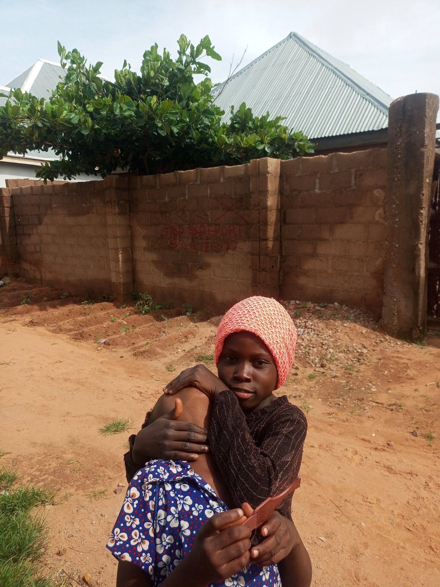 In the city of Tempure, Nigeria, two children are photographed outdoors. One child, positioned in the foreground, wears a pink knitted head covering and a dark, shimmering long-sleeved shirt. This child has their arms around another child, whose head is not visible, but who is wearing a blue dress with a white floral pattern. The child in the foreground is holding a reddish-brown object, possibly a comb, in their right hand. The background features a brown brick wall with red graffiti. Beyond the wall, a tree with lush green leaves is visible, along with a silver, corrugated metal roof of a building. The ground is sandy, with patches of dry grass. The sky above is overcast.
