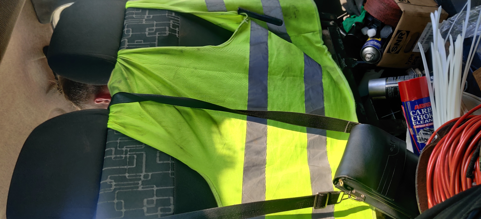 The image captures the cluttered interior of a vehicle, likely a car or van, seen from a high angle. A person is lying across the vehicle's seat on the left, with only the back of their head and an ear visible. They are wearing black attire.
A bright fluorescent yellow safety vest with grey reflective stripes is draped over the seat and the person, covering much of the frame's center. A black strap, possibly from a bag or a seatbelt, crosses over the vest. Towards the bottom right, a black leather-like bag or case with metal buckles is visible.
To the right of the safety vest, various items are scattered, suggesting a workspace or storage area. These include several aerosol cans: a blue and white can faintly showing "WD-40", a plain silver can, and a prominent red and blue can labeled "ABRO CARB CHOKE CLEAN" with "LIMPIADOR DE CARBURADORES" underneath. A cardboard box with "SNR" printed on it is partially visible. A pile of white cable ties, a coiled red electrical wire or cable, a small black brush-like tool, and a green cloth are also among the items.
The scene is brightly lit by natural light, suggesting it is daytime. No specific activity is occurring other than the person resting amidst these various tools and work-related items. The seat upholstery has a dark pattern resembling circuit board lines.
