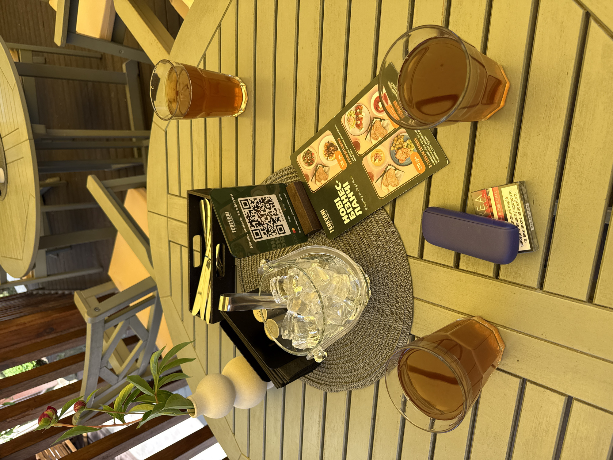 An outdoor table at a restaurant in Vinnytsia, Ukraine, is set with two glasses of amber-colored liquid, likely iced tea or a similar beverage. An ice bucket filled with ice, a salt and pepper shaker set, and silverware in a black holder are also present on the table. A small vase with a flowering plant sits to the left, and a folded menu with food images is in the foreground. A QR code printed on a stand and a pack of cigarettes are also visible. The table surface is made of weathered, pale yellow wooden planks. In the background, chairs and other tables are visible, along with wooden fencing and greenery. The scene appears to be a leisurely outdoor dining setting.