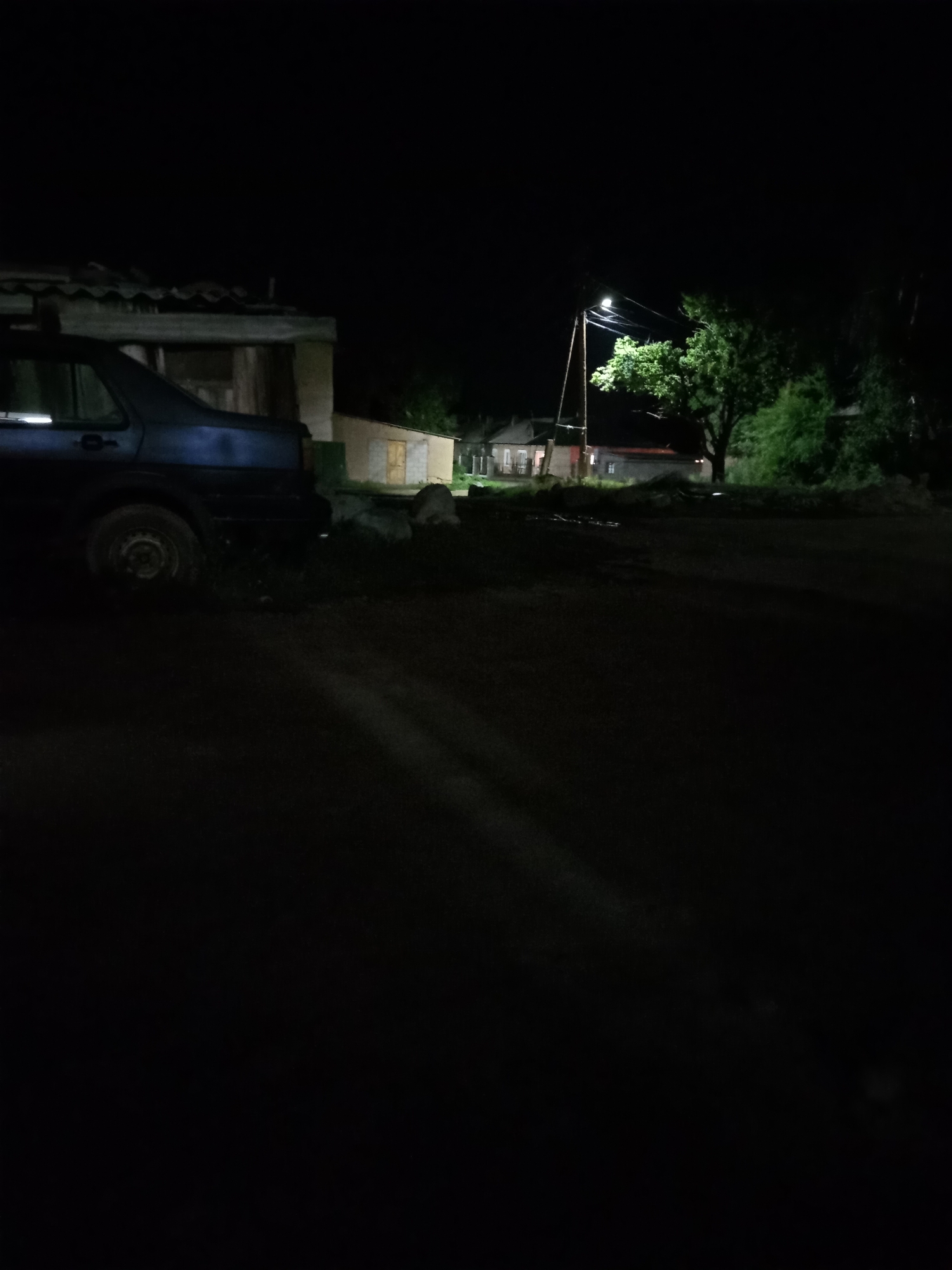 This is a nighttime photograph taken in Burma-Suu, Kyrgyzstan. The scene depicts a dimly lit rural area with a few buildings and a car. The foreground is dark, with shadows cast across a dirt road. On the left, the rear of a dark-colored car is visible, parked next to a dilapidated structure with a corrugated roof. Further into the scene, a streetlamp illuminates a large, leafy tree. Behind the tree, several houses are visible, appearing as simple, possibly residential structures. The overall atmosphere is quiet and still, suggesting a peaceful rural evening. There is no visible text in the image.