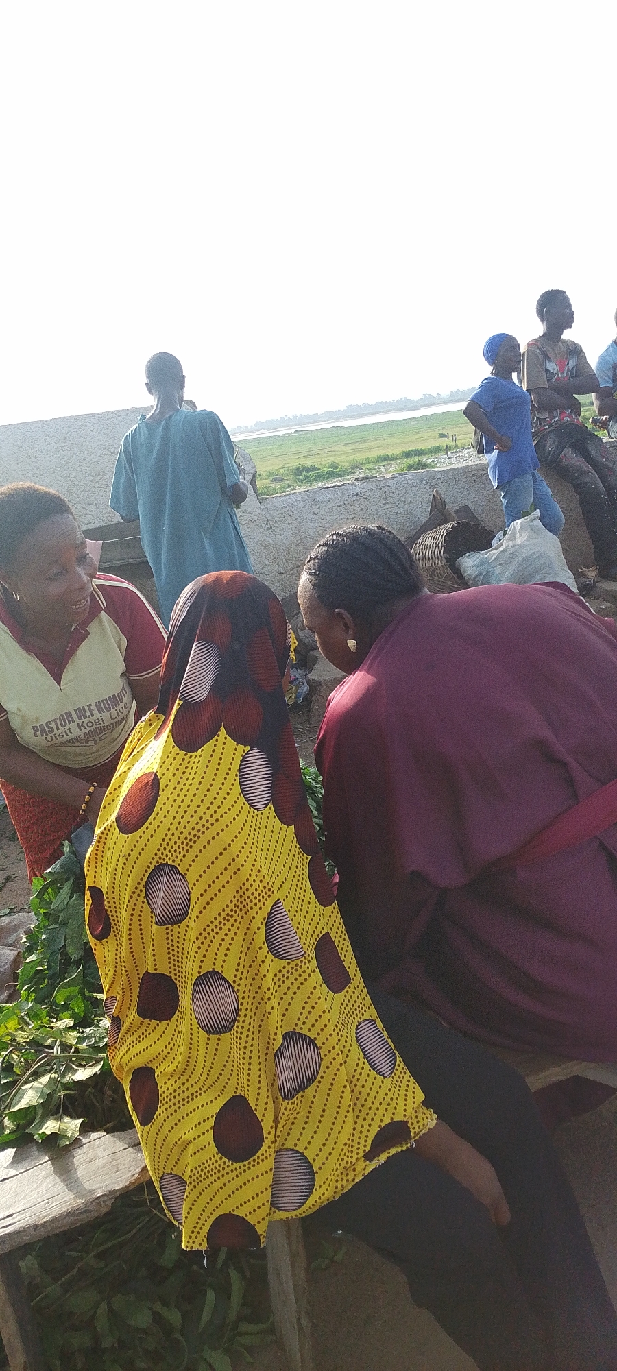 In Lokoja, Nigeria, a group of people are gathered in an outdoor setting. A woman wearing a yellow patterned headscarf and a matching yellow top with brown circles is seated. To her left is a woman wearing a light yellow t-shirt with red sleeves and lettering. To the woman's right is another woman wearing a dark red top. They appear to be in conversation. Behind them, several other people stand near a low wall that overlooks a grassy area and a body of water. The background suggests a rural or semi-rural environment.