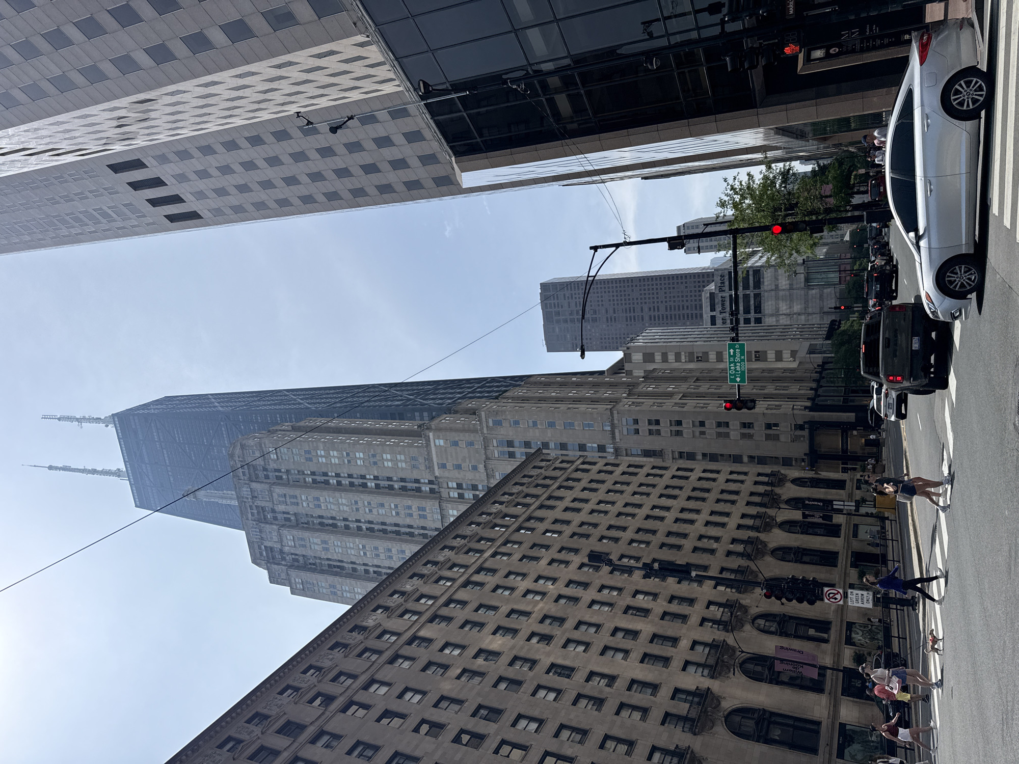 This media file depicts a street scene in Chicago, USA, looking upwards towards tall buildings. The sky is clear and bright, suggesting it is daytime. Several people are visible, some crossing a street at a crosswalk, while others appear to be walking along the sidewalk. A silver car is parked at the crosswalk, and other vehicles are present in the background, indicating moderate traffic.
The foreground is dominated by the lower portions of several buildings with distinct architectural styles. One building features a repeating pattern of arched windows and ornate detailing, while another appears more modern with a grid-like facade. In the background, a very tall, dark-colored skyscraper with a distinctive silhouette, likely the John Hancock Center, is prominent. Other modern skyscrapers are also visible, showcasing Chicago's characteristic urban landscape.
Several traffic lights are visible, one showing a red light. Street signs are also present, including one indicating "E Oak St" and "Lake Shore Dr 1000 N." Banners or signs on one of the buildings read "Drawing" and "William Kooning." The overall impression is that of a busy urban environment on a clear day, with a mix of historic and modern architecture. The perspective from below emphasizes the grandeur and scale of the city.