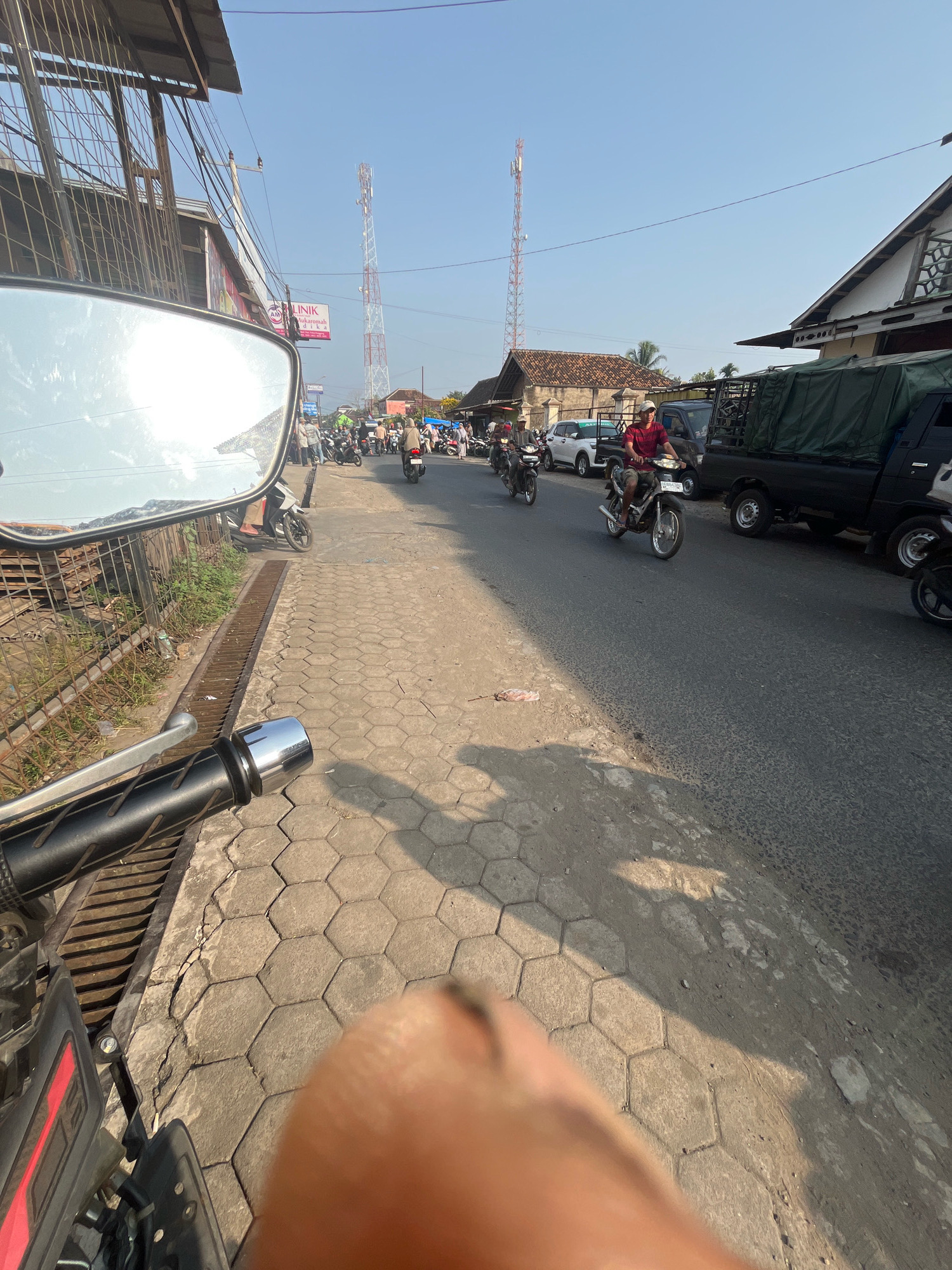 The image depicts a street scene in Kemuning, Indonesia, viewed from the perspective of someone riding a motorcycle. In the foreground, the handlebar and part of a motorcycle are visible, along with a side-view mirror reflecting the street. The street is paved with hexagonal concrete blocks on the left side and asphalt on the right. A metal grate runs along the edge of the sidewalk.
The street is moderately busy with traffic. Several motorcycles, some with riders, are moving along the asphalt road. One motorcycle in the mid-ground has a rider wearing a red shirt and a white cap. Further down the road, a black truck with a green tarp covering its cargo is parked. Buildings line the street on both sides, with some appearing to be shops or houses. A tall telecommunications tower is visible in the distance against a clear blue sky.
The overall environment suggests a typical urban or semi-urban setting in Indonesia during daylight hours. The presence of multiple motorcycles, the style of the buildings, and the telecommunications tower are characteristic of such locations.
