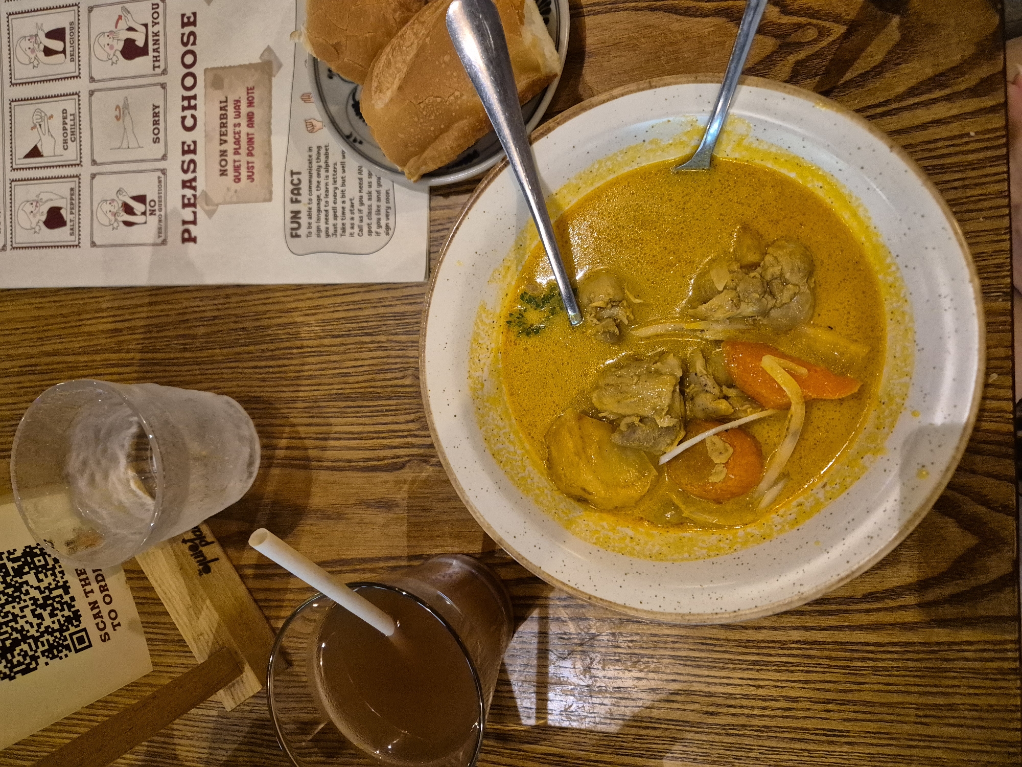 This is an overhead, close-up shot of a meal on a wooden table. The meal consists of a bowl of yellow curry with pieces of chicken and carrots, accompanied by a glass of brown liquid with a straw, and a plate of bread rolls. To the left of the meal, a QR code is displayed on a stand, with text that reads "SCAN THE TO ORDI". Next to the QR code is a glass of water. In the background, a chart displays various hand signals with descriptions such as "SALT, PEPPER", "CHOPPED CHILLI", "DELICIOUS", "SORRY", and "THANK YOU". Below this chart, the text "PLEASE CHOOSE" is prominently displayed, with a smaller note below it stating "NON VERBAL QUIET PLACE'S WAY. JUST POINT AND NOTE." A "FUN FACT" section at the bottom of the chart discusses learning sign language. The overall setting suggests a restaurant environment. The city of Đà Nẵng, Vietnam, is not explicitly mentioned or depicted in the image.