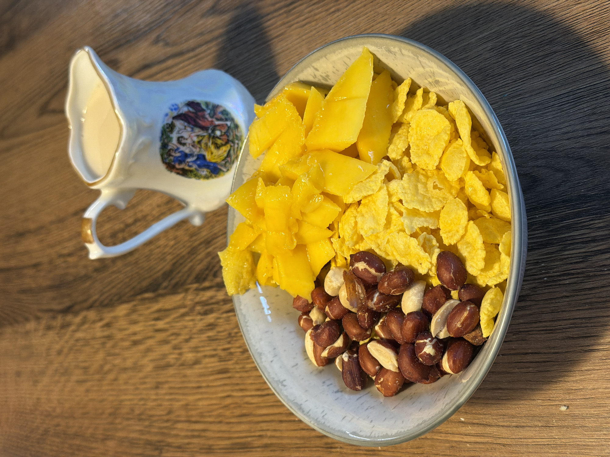 A bowl of cereal, mango slices, and nuts sits on a wooden table. To the right of the bowl is a small pitcher filled with a light-colored liquid, possibly milk or cream. The pitcher is white with a gold rim and features a decorative image of people. The table is made of wood with a visible grain pattern. The lighting suggests an indoor environment, with a shadow cast by the pitcher onto the table. There are no people visible in the image. The location in the city of Perm, Russia, is not discernible from the image.