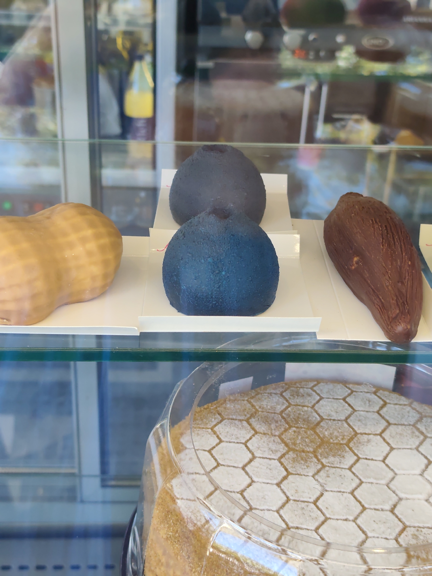 The image shows a display case in what appears to be a bakery or confectionery shop in Sochi, Russia. In the upper section of the display, several pastries are visible. On the left, there is a light brown pastry shaped like a peanut. To its right, there are two spherical, dark blue pastries, one slightly larger and positioned behind the other. Further to the right, there is a dark brown, elongated pastry resembling a cocoa pod or a large bean.
In the lower section of the display, a cake is visible under a clear plastic dome. The cake's top is decorated with a honeycomb pattern formed by powdered sugar and possibly a light brown glaze. The overall environment suggests a commercial food establishment with glass display cases. No people are visible in the image.