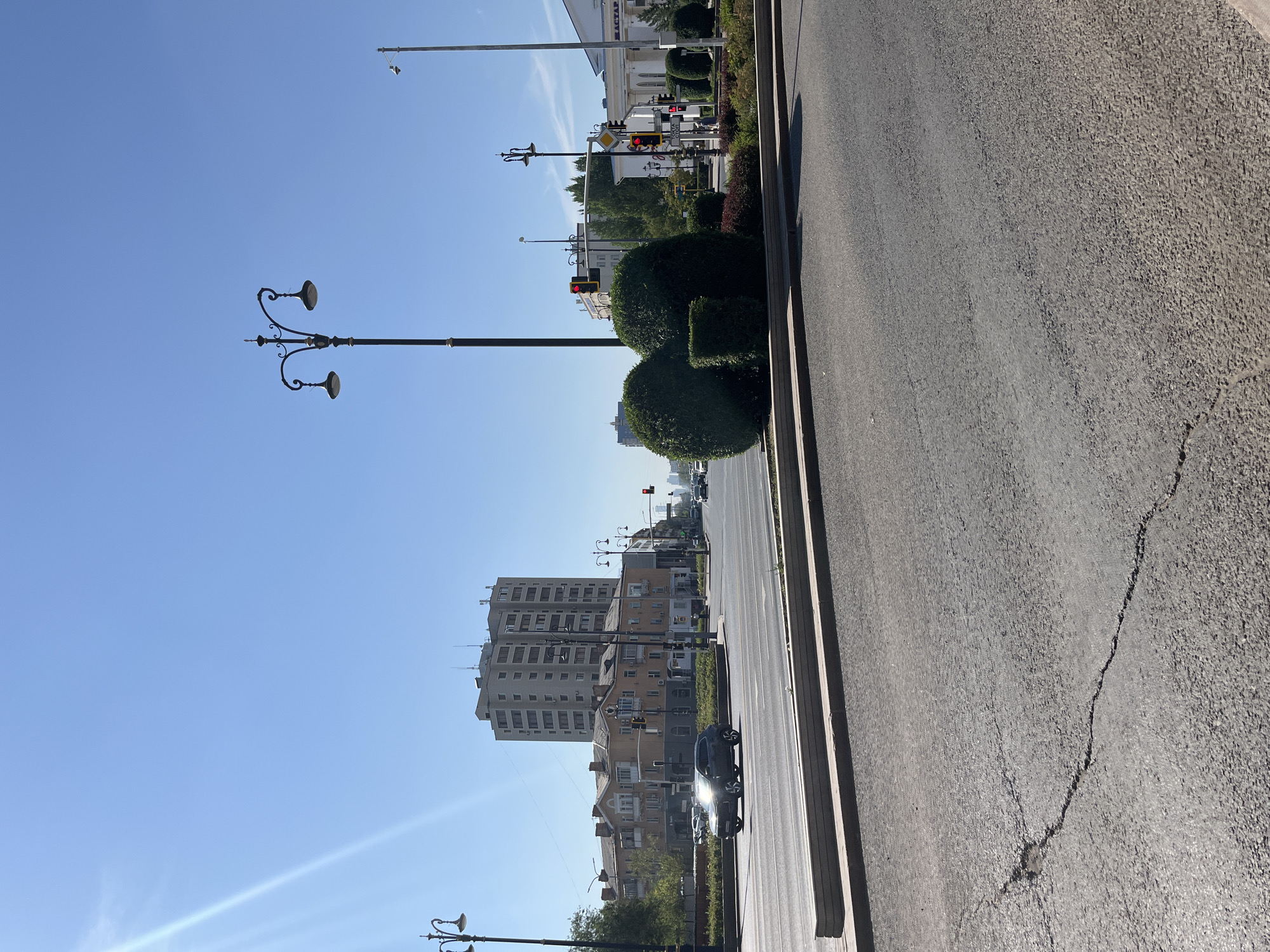 This image depicts a street scene in Astana, Kazakhstan, likely on a sunny day. The foreground is dominated by a cracked asphalt road with a concrete median strip. In the middle ground, a multi-lane road stretches across the frame, with a dark-colored SUV driving on it. Beyond the road, there are several buildings, including a tall, modern apartment building and a lower, older residential building with balconies. Decorative bushes and trees are visible along the street. Multiple lampposts with ornate designs are present, as are traffic lights and traffic signs. In the background, more buildings and trees are seen under a clear blue sky, with a faint lens flare visible in the upper left corner. The overall impression is of an urban environment with a mix of old and new architecture.
