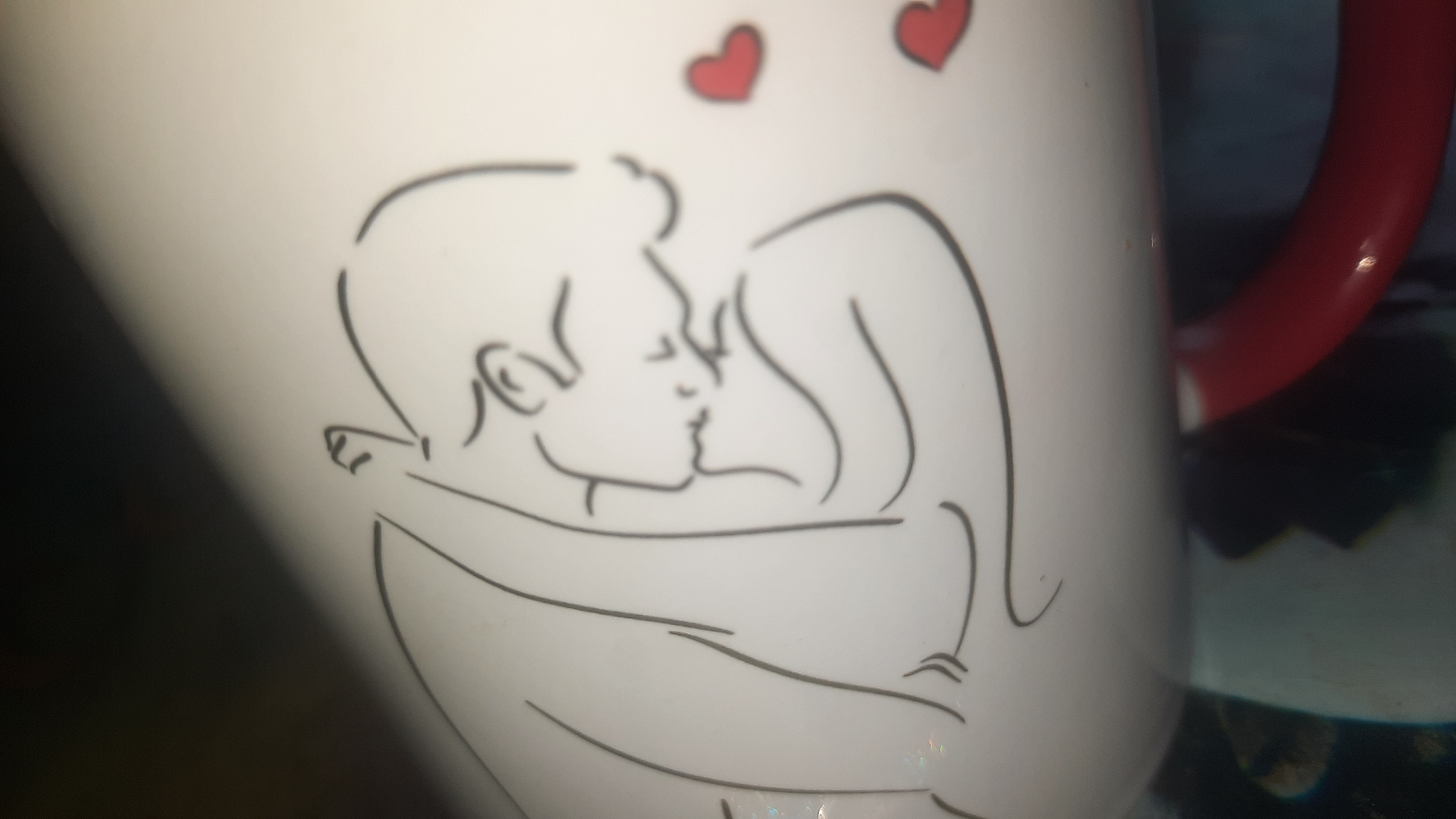 The image displays a close-up of a white mug with a red handle. The mug features a black line drawing of a couple embracing and kissing, with two small red hearts above their heads. The background is blurred, showing a dark, indistinct environment that may be an indoor setting. There is no discernible information about the location of Shklov, Belarus.