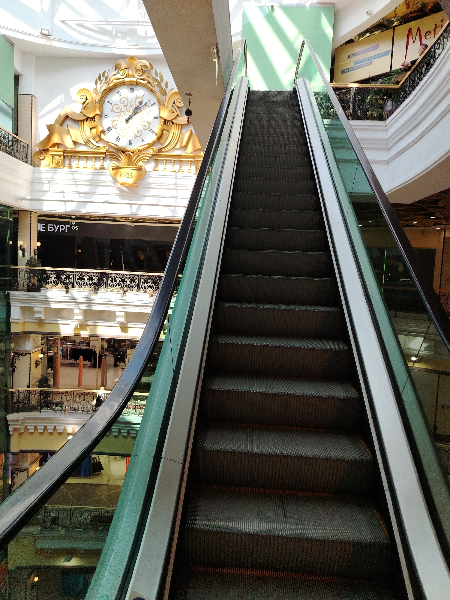 The image depicts the interior of a multi-level shopping mall, with a prominent escalator occupying the foreground. The escalator is angled upwards and to the right, with dark gray steps and white handrails on either side, bordered by glass panels.
In the background, on the upper level of the mall, a large, ornate golden clock is mounted on a white wall. The clock face features Roman numerals and decorative golden elements. Below the clock, the year "2016" is visible.
Further back and to the right, a large sign with the word "Metropolis" in red lettering is visible on a light-colored background, with a person standing on a balcony to its right.
The mall exhibits several levels, with balconies featuring decorative metal railings and planters with greenery. Shops are visible on the lower levels, with clothing racks and displays. Sunlight streams in from the ceiling, creating shadows and illuminating parts of the interior. The overall environment suggests a bright and busy retail space.
