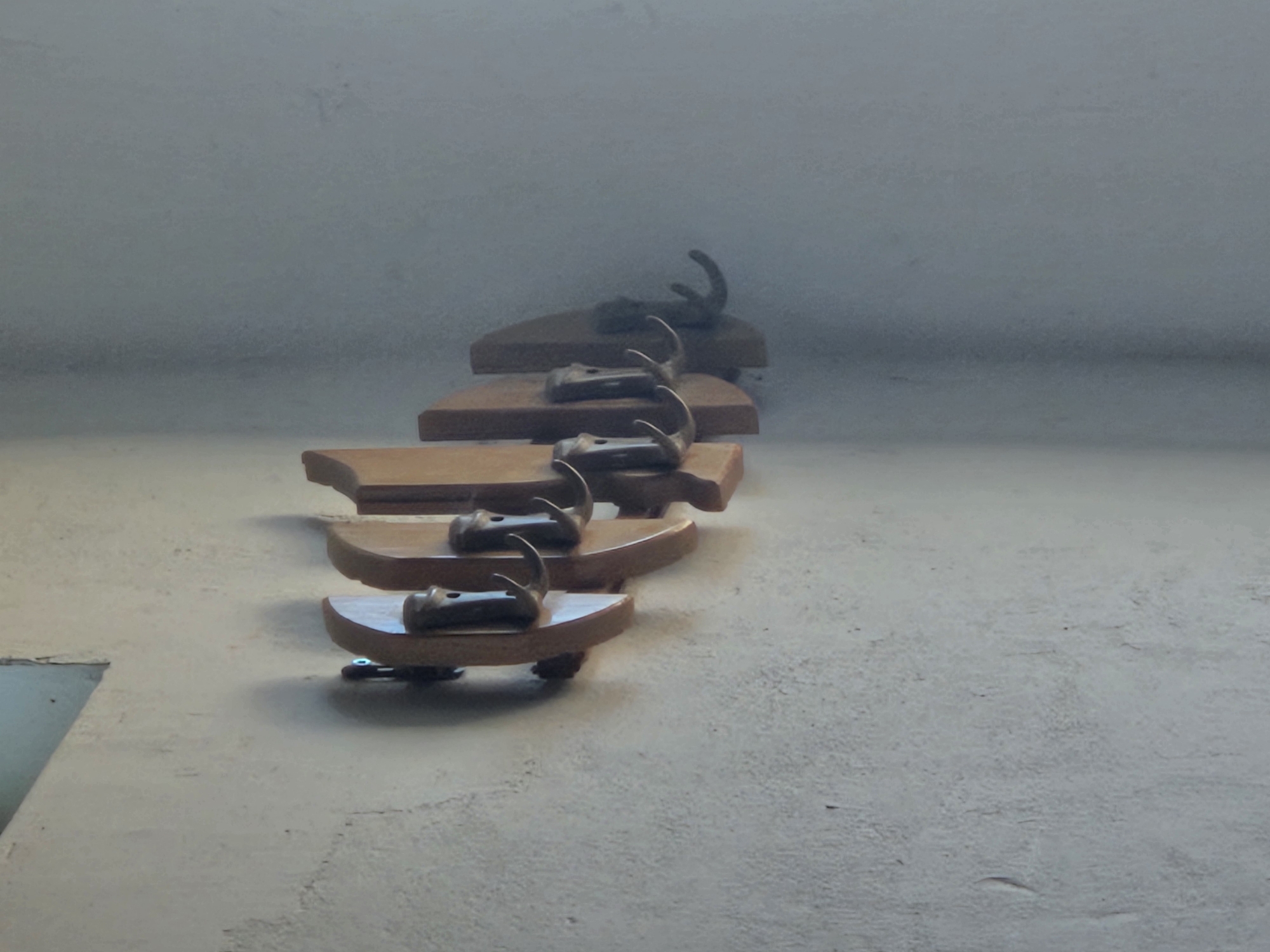 A wooden coat rack with five hooks is mounted on a textured, light-colored wall. The coat rack is composed of individual wooden pieces, each housing a metal hook. The hooks are curved and appear to be made of a dark metal. The wall has a rough, uneven surface, possibly plaster or stucco, with some visible imperfections and shadows. To the left of the coat rack, a small section of a light blue or grey rectangular object is visible, possibly a window or a framed picture. The lighting suggests an indoor environment, with shadows cast by the coat rack and hooks onto the wall.