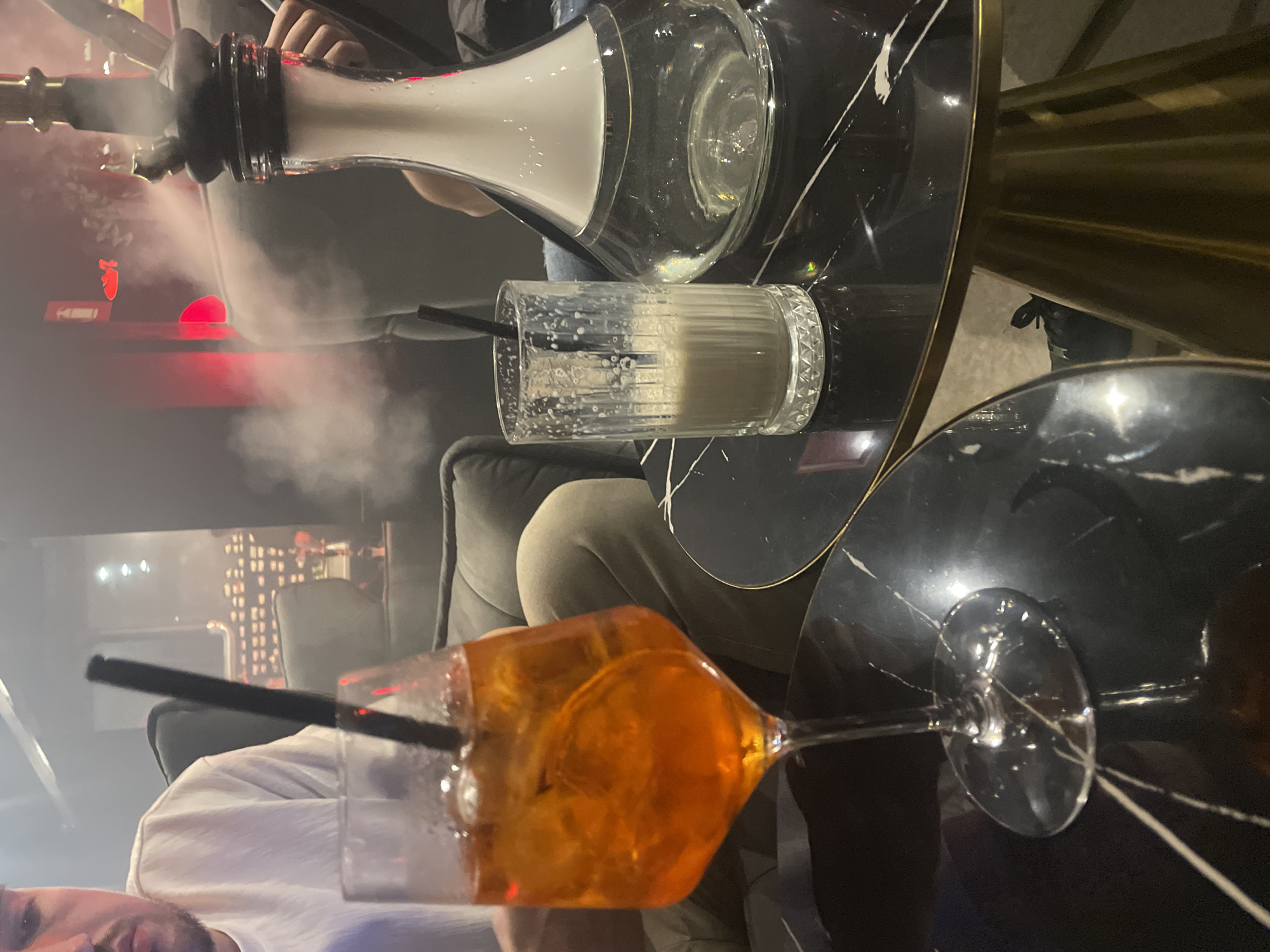A dimly lit interior setting appears to be a lounge or bar, potentially in Varna, Bulgaria. Smoke hangs in the air, possibly from a hookah visible in the upper left, held by an unseen person. On a black marble-patterned table, two glasses are situated. One contains an orange-colored cocktail, garnished with an orange slice and a black straw. Another glass holds a lighter-colored beverage, also with a black straw. A partial view of a seated individual in light-colored clothing is present. In the background, blurred lights suggest an urban setting with illuminated buildings.