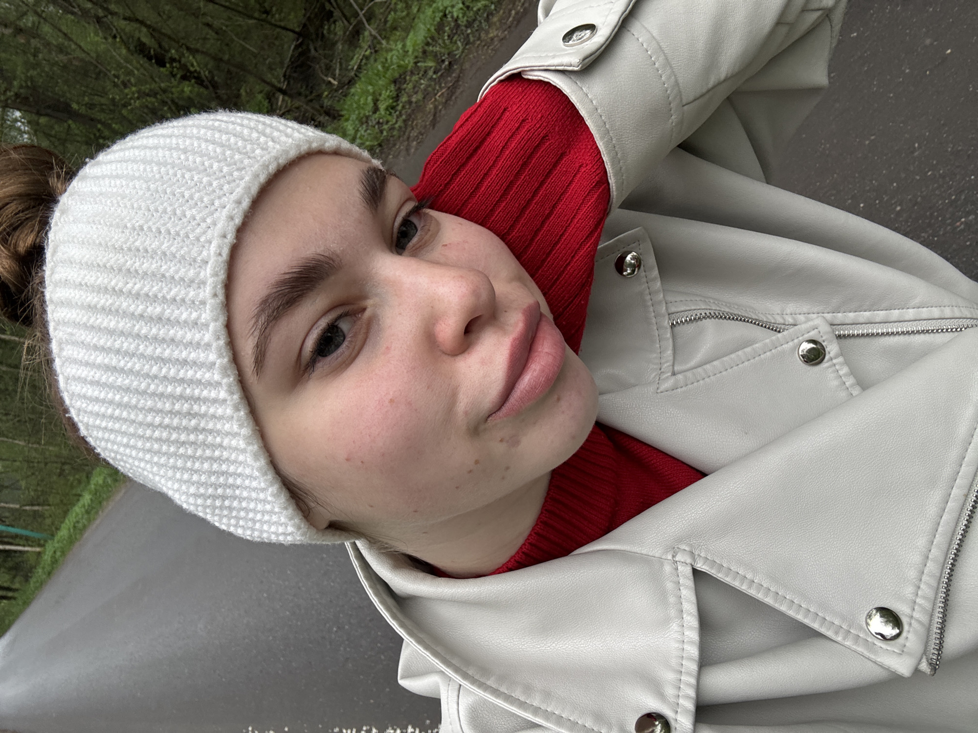 A selfie of a woman with fair skin, brown hair pulled back in a bun, wearing a white knit cap, a red ribbed sweater, and a white leather jacket. She is in an outdoor setting, likely on a paved road or path with greenery visible in the background. The location is identified as Мещёры, Russia.