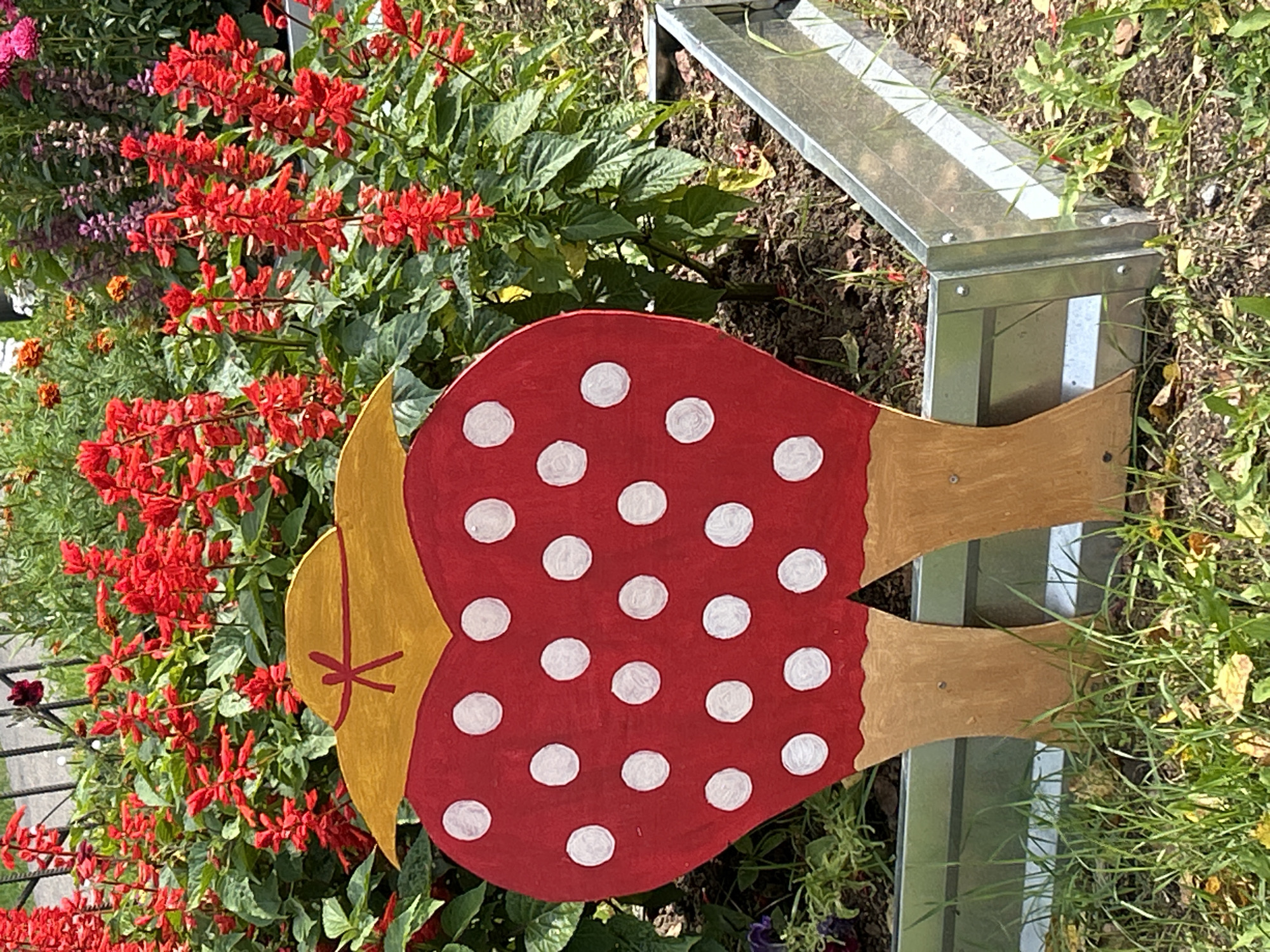 This is a photograph taken outdoors, likely during daylight hours, in a garden setting. The foreground features a cutout decoration that resembles a large mushroom, painted red with white polka dots. The mushroom has a yellow-painted hat with a red ribbon and a stem that is divided into two legs, painted brown. The mushroom decoration is positioned in front of a flower bed filled with vibrant red flowers and lush green foliage. A metal garden border is visible to the right, separating the garden bed from the surrounding soil and grass. The overall impression is one of a decorative garden element situated amongst blooming plants. The location is described as being in the city of Tomsk, Russia.